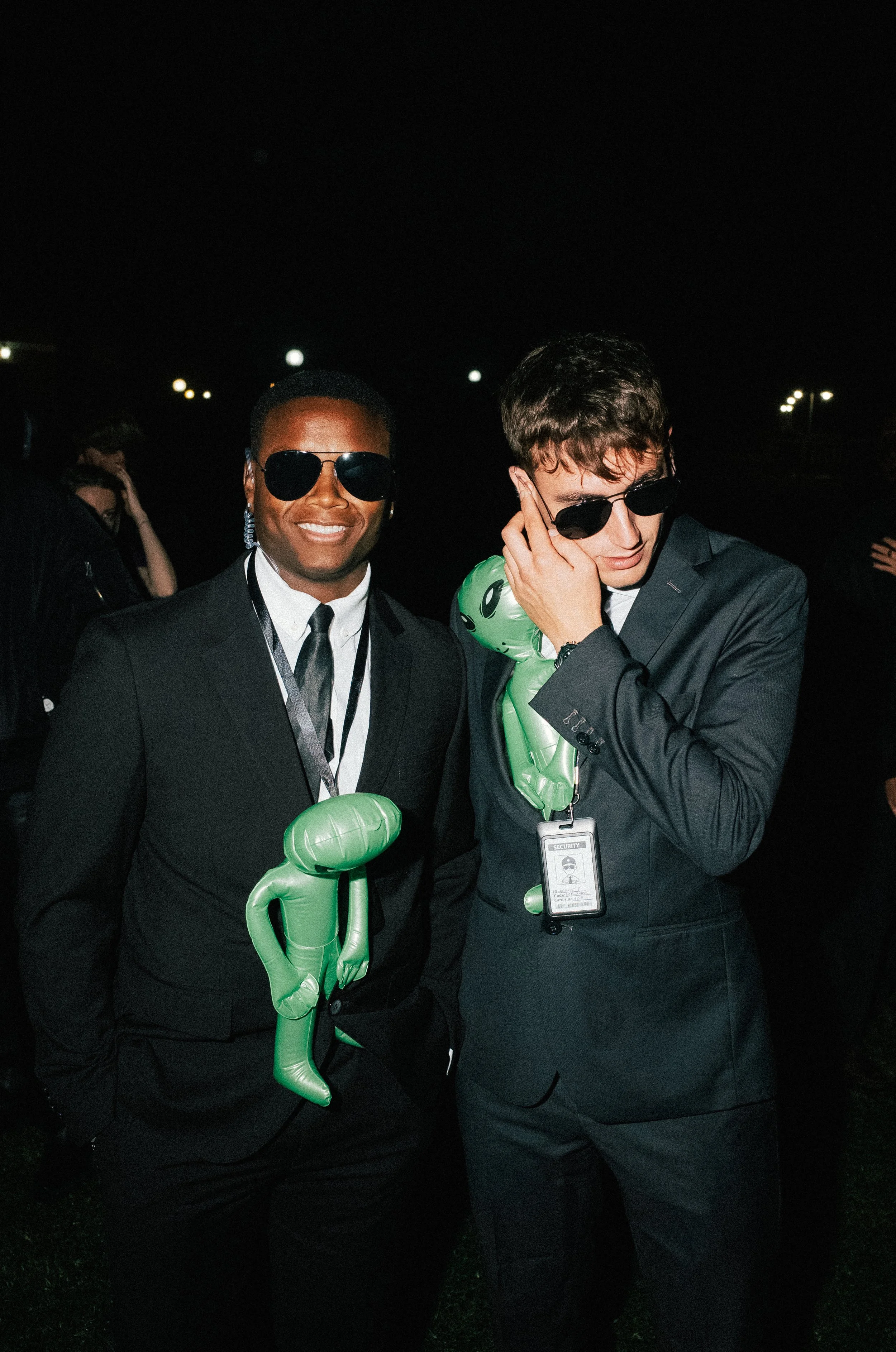 Two men in black suits and sunglasses at night, each with a green inflatable alien around their necks, standing outdoors.
