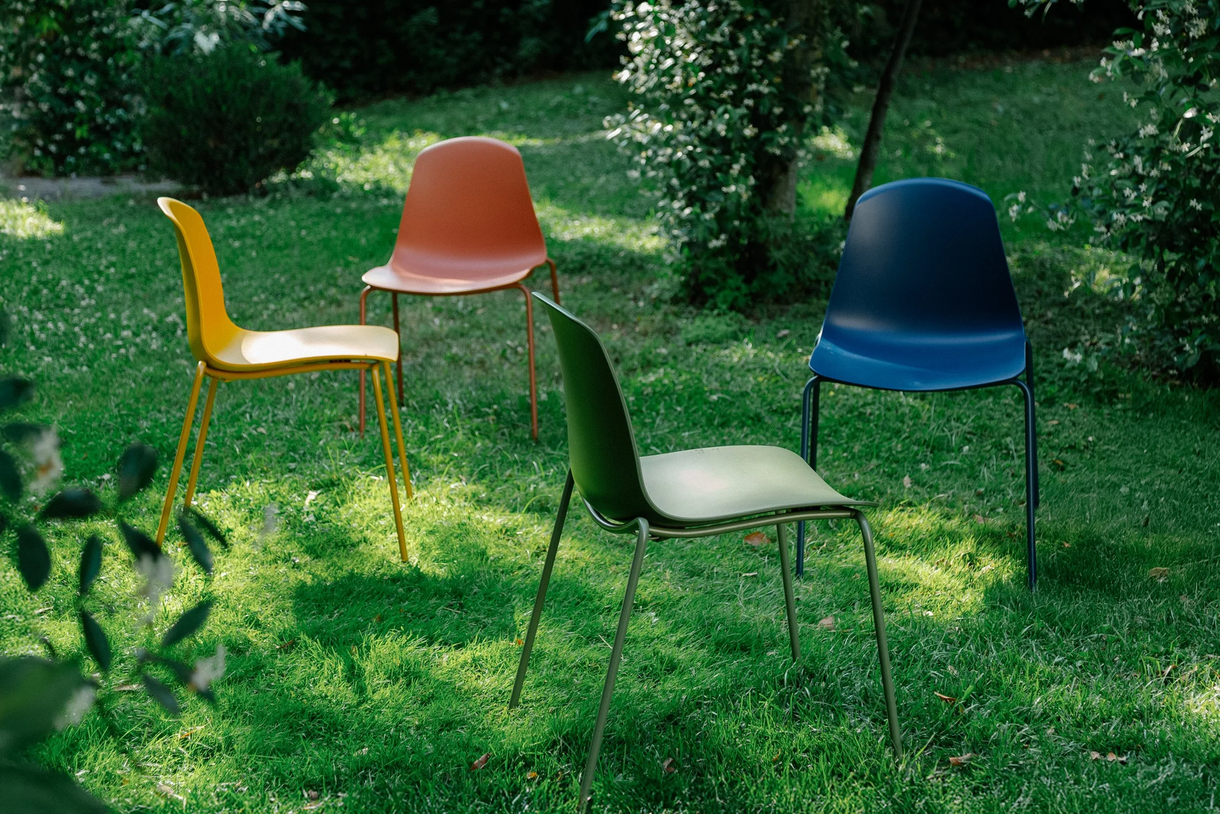 Four colorful chairs in a grassy outdoor garden, surrounded by green bushes and trees.