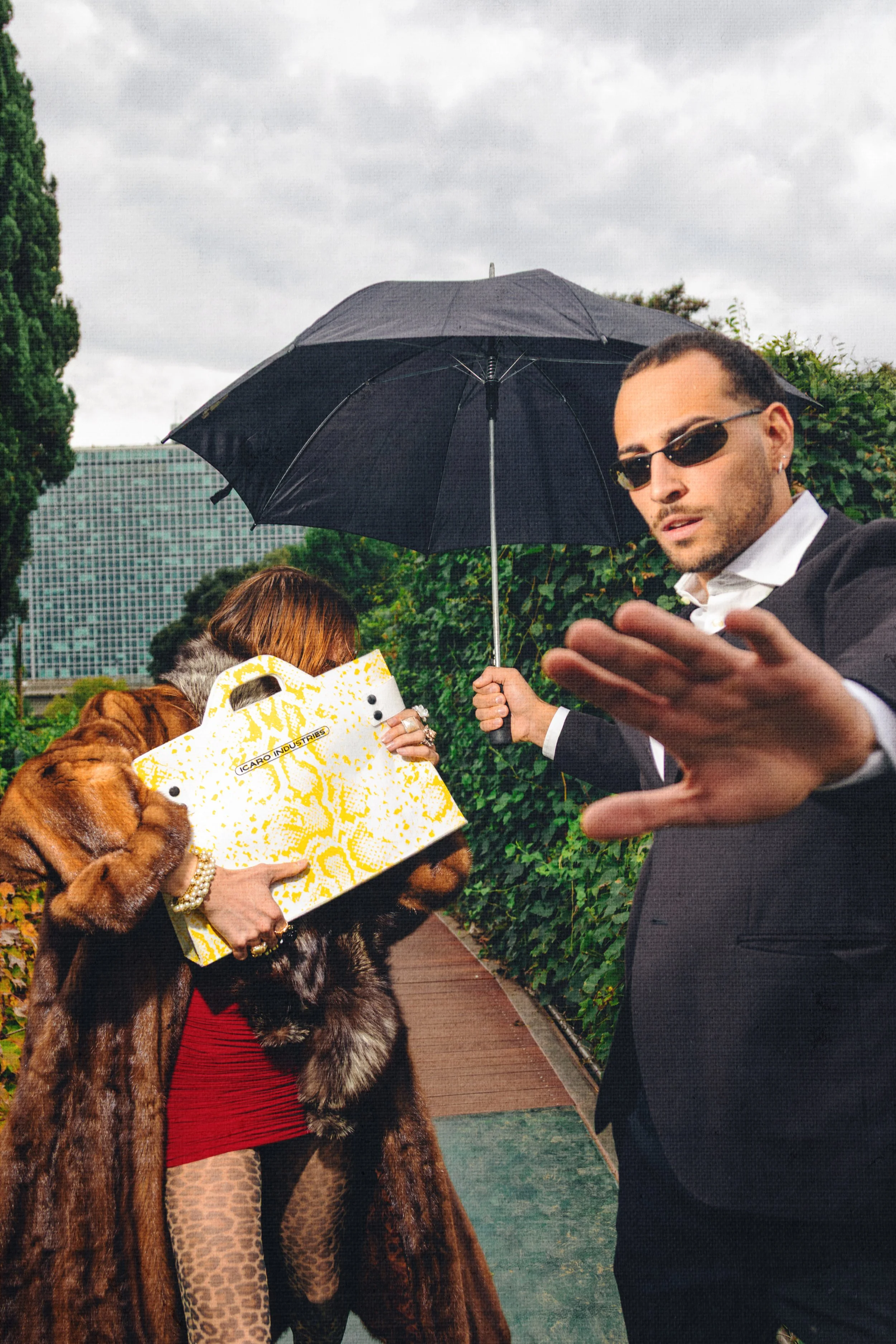 A man in sunglasses and a suit is holding an umbrella, speaking towards a woman who is carrying a luxury shopping bag, standing outdoors on a sidewalk with green bushes and tall buildings in the background.