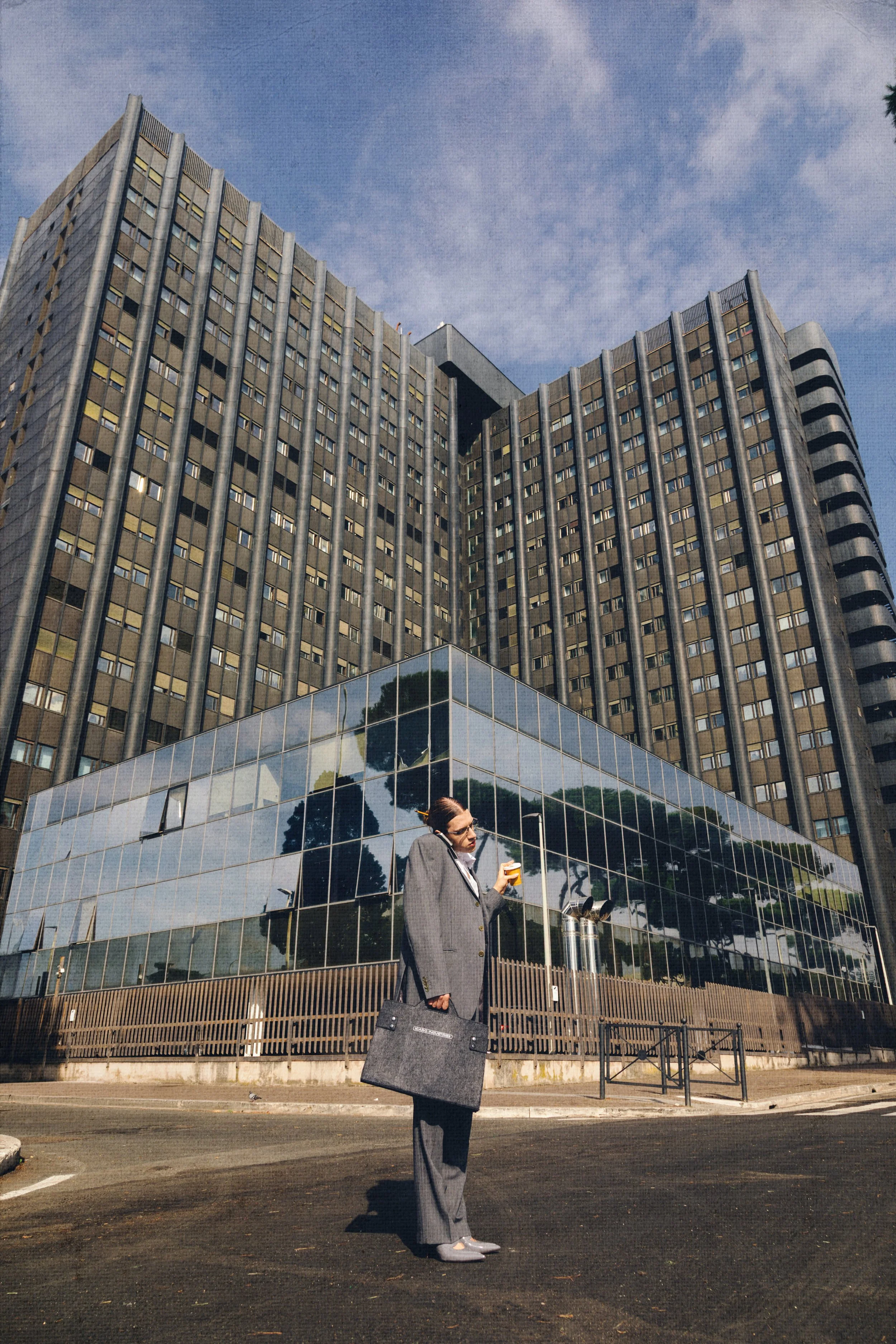 A woman in a gray business suit stands on a city street holding a drink and a briefcase, with tall modern office buildings reflected in glass windows behind her on a sunny day.