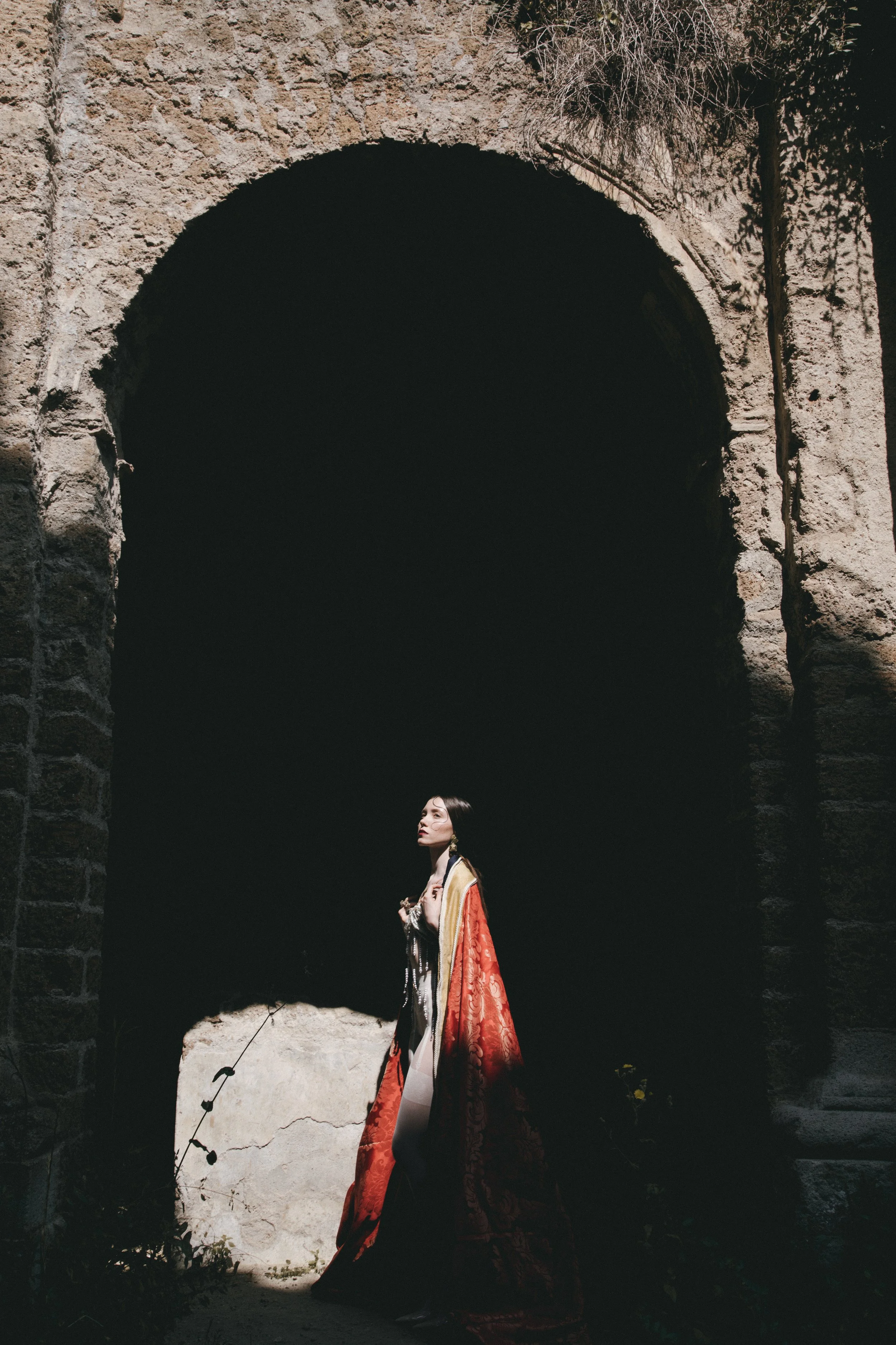 A woman wearing an elaborate, colorful coat standing beneath a large stone archway, with dramatic lighting highlighting her against the dark background.
