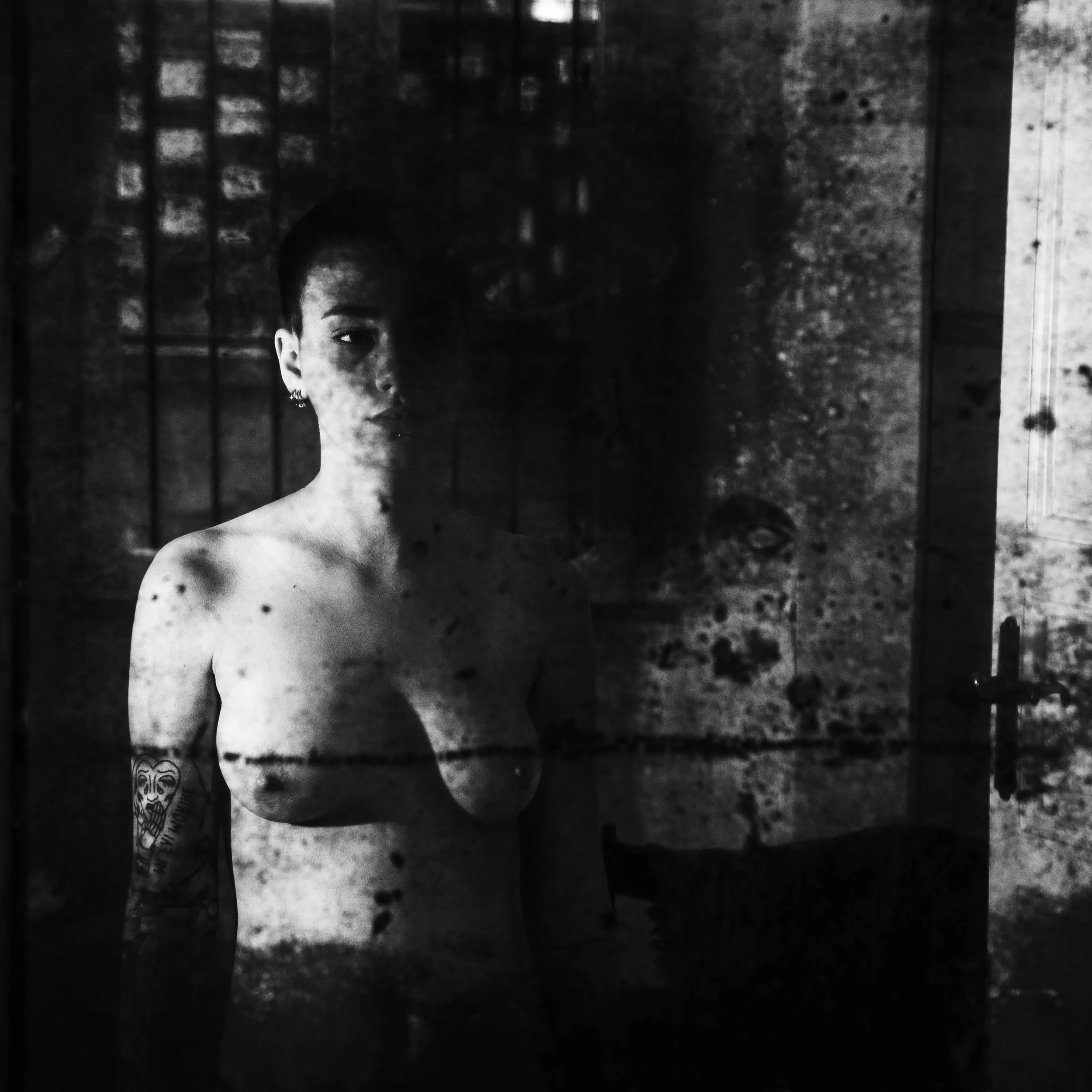 A black and white photograph of a woman standing behind a dirty, scratched glass door with a reflection partially obscuring her face. She has short hair, earrings, and a tattoo on her left arm.