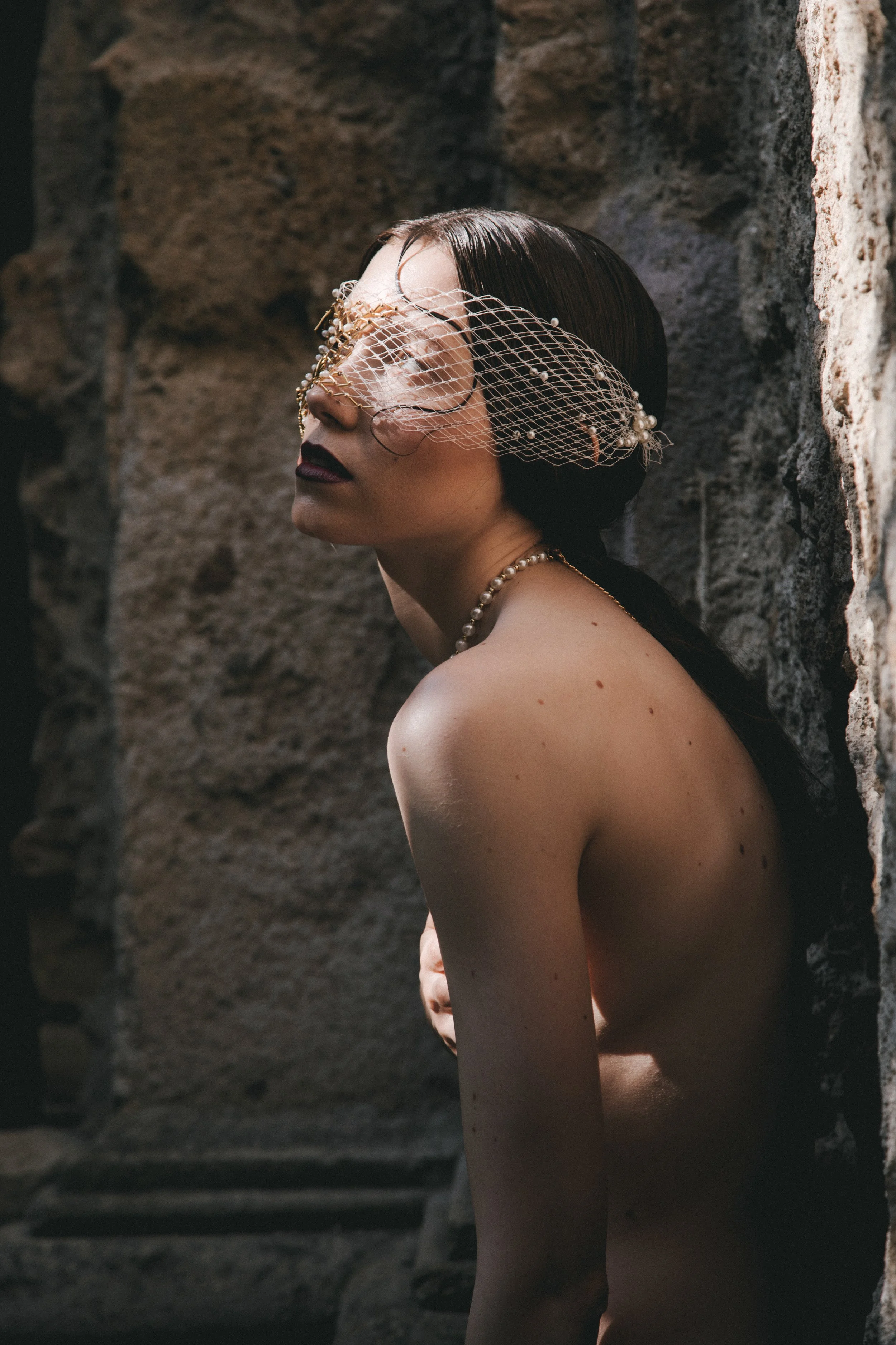 A woman with dark hair and dark lipstick, wearing a pearl necklace and a decorative face net, is leaning against a stone wall with her eyes closed.