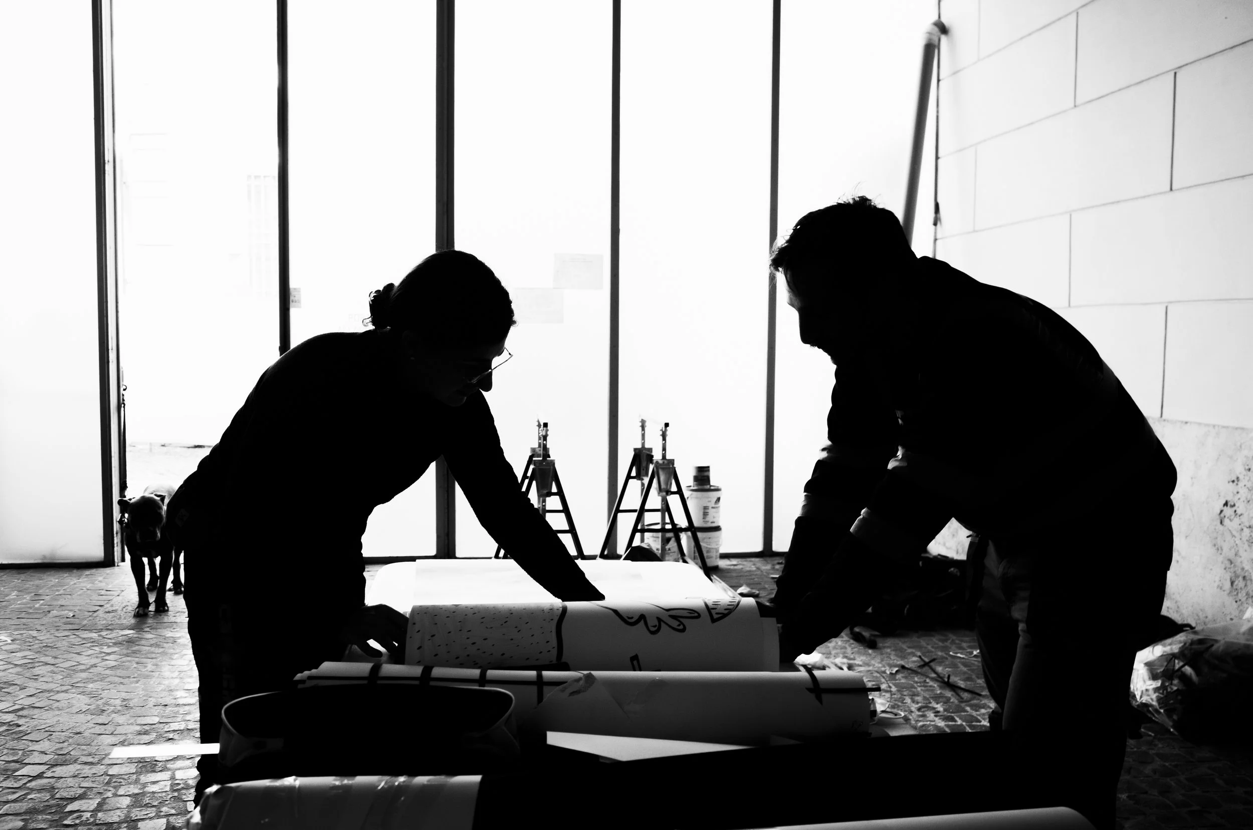 Silhouettes of two people working on a project with rolls of paper or plans on a table in a bright indoor space, with a dog in the background.
