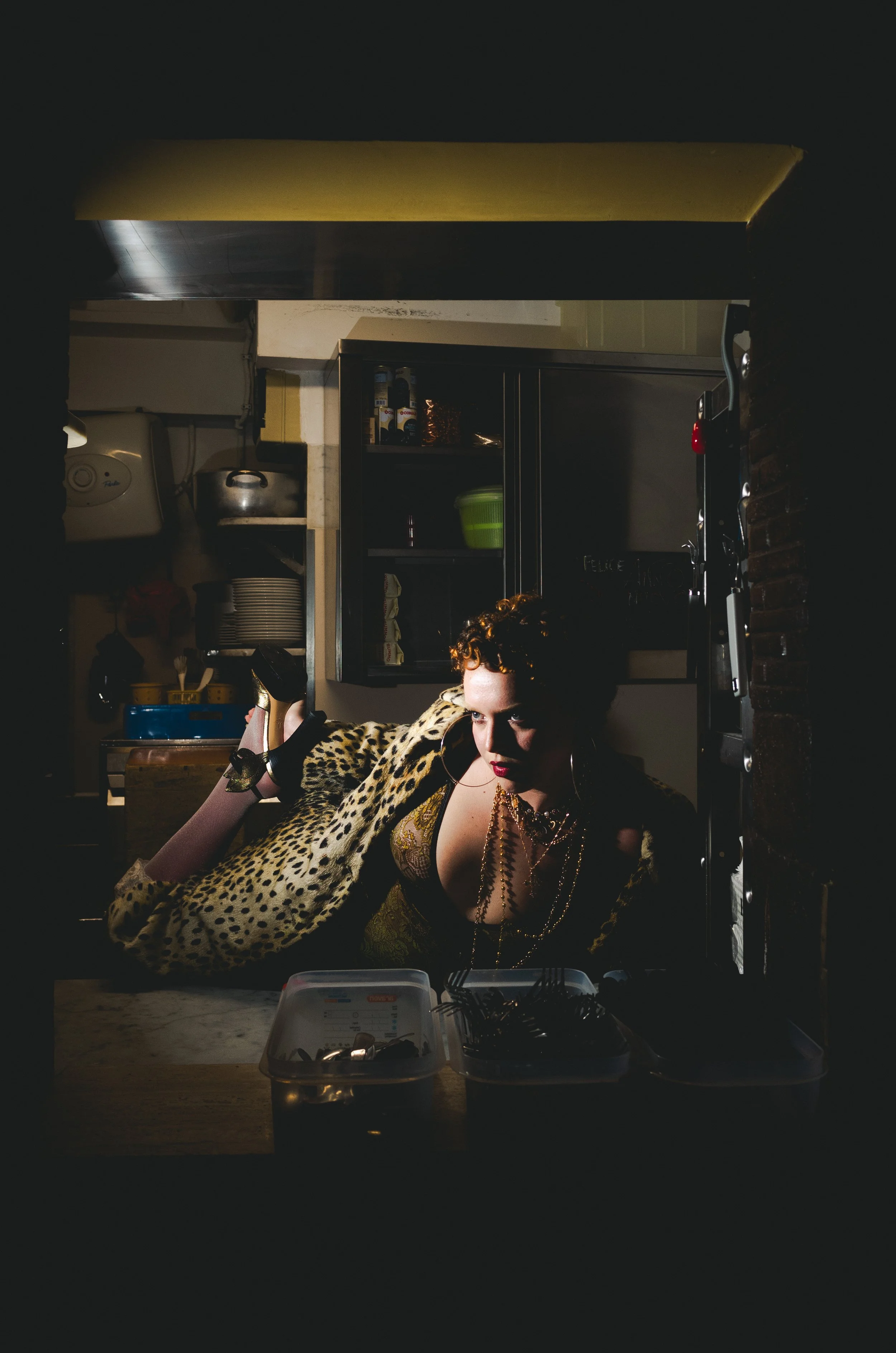 A woman with curly hair wearing a leopard print jacket and gold necklace lying on a kitchen counter, illuminated by a small light, with kitchen shelves and utensils in the background.