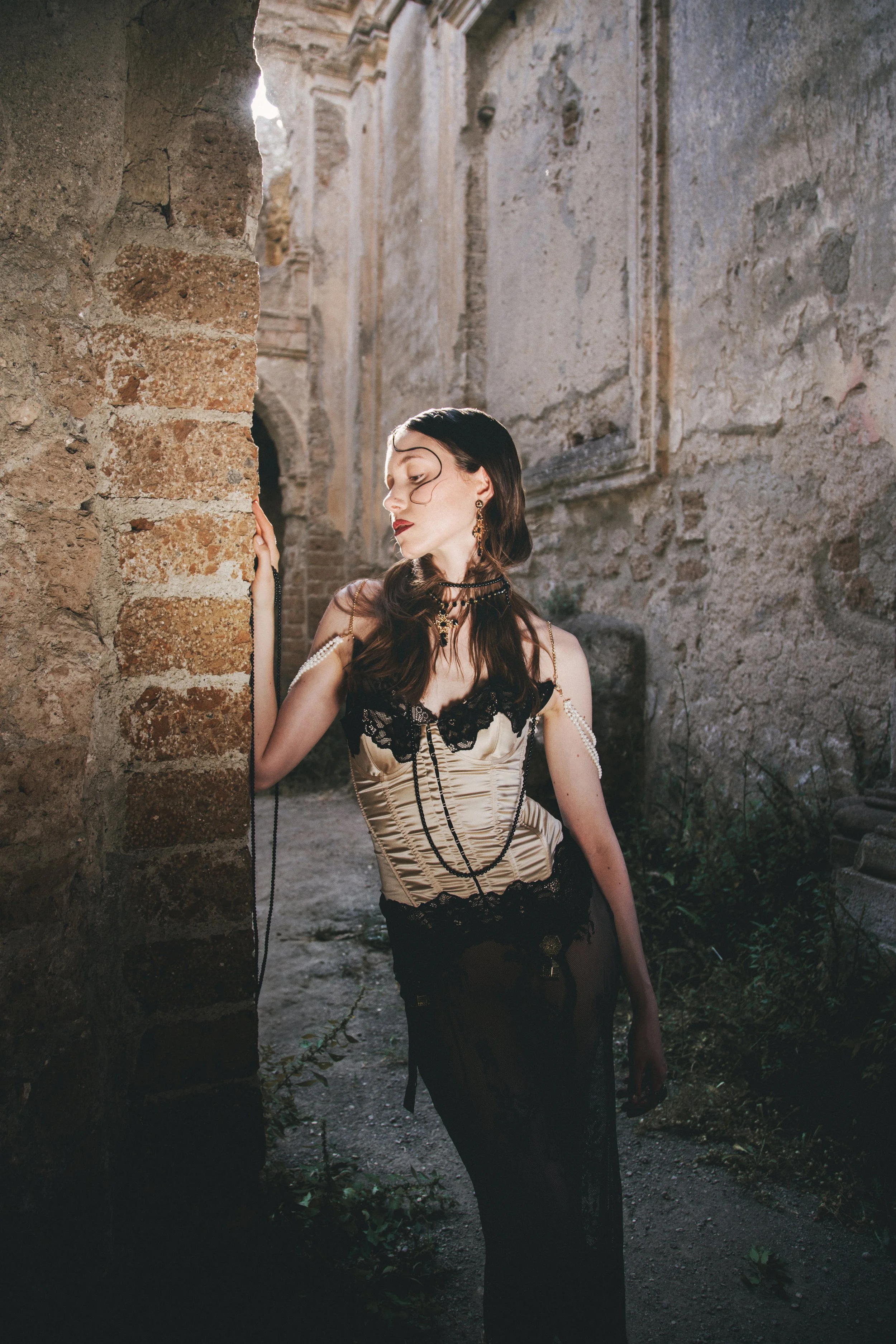 A woman in vintage-style clothing and jewelry stands against an ancient stone ruin, with ruins and steps visible in the background.