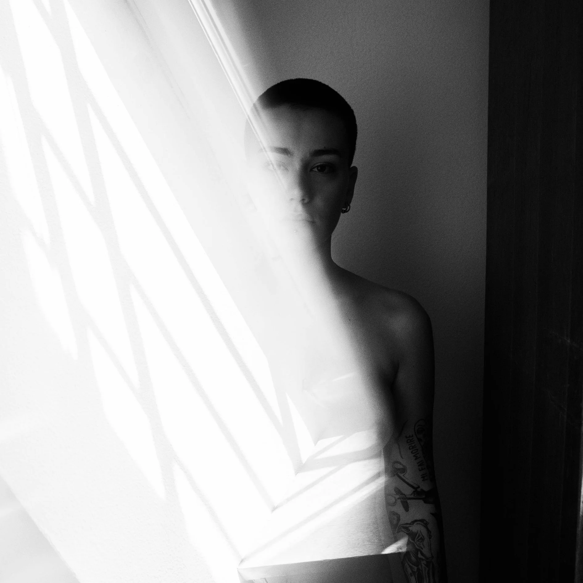 Black and white photo of a woman with short hair and tattoos, partially obscured by a frosted glass door.