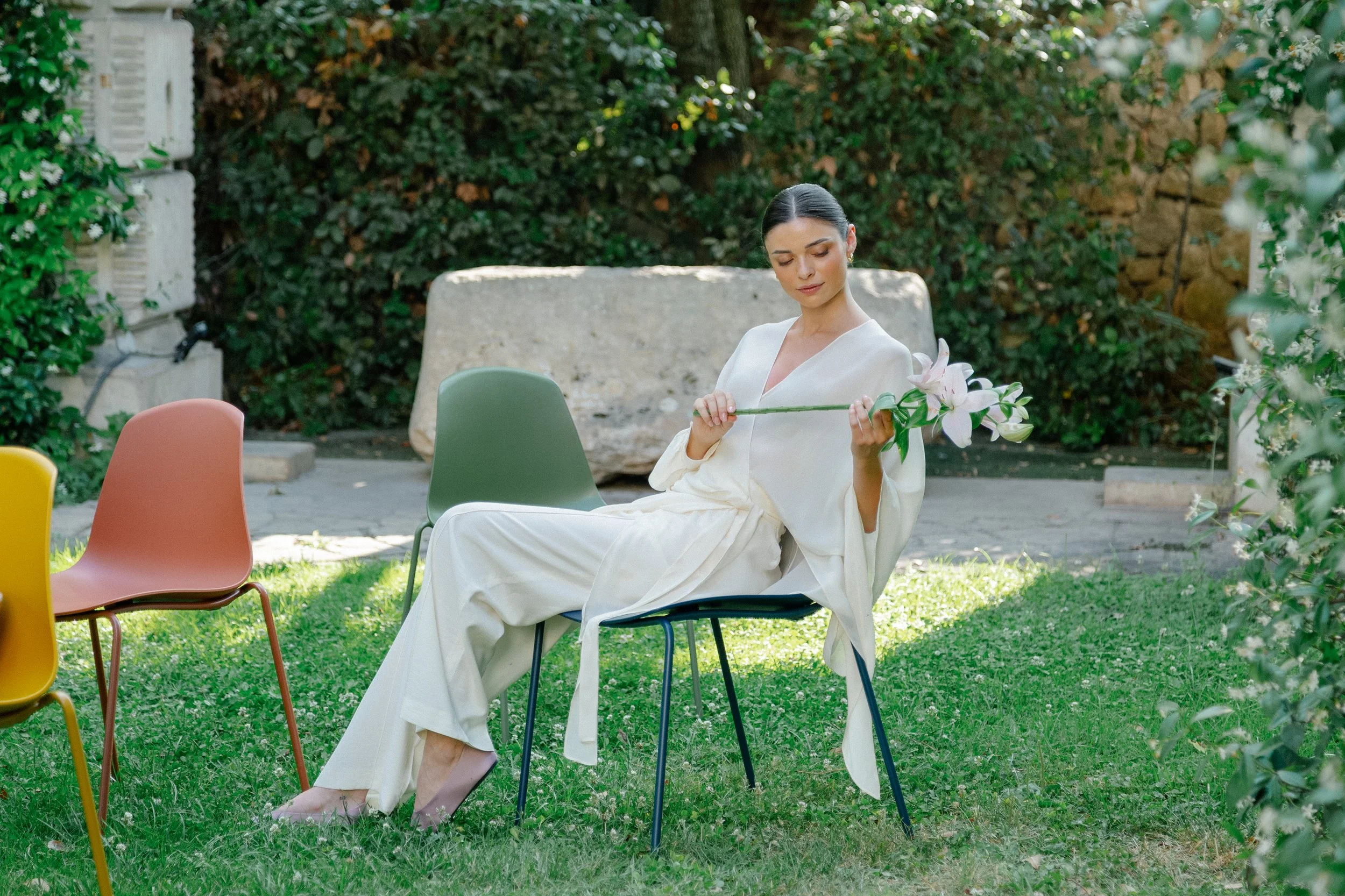 A woman dressed in white sitting on a chair in a garden, holding a white lily flower, surrounded by colored chairs and greenery.