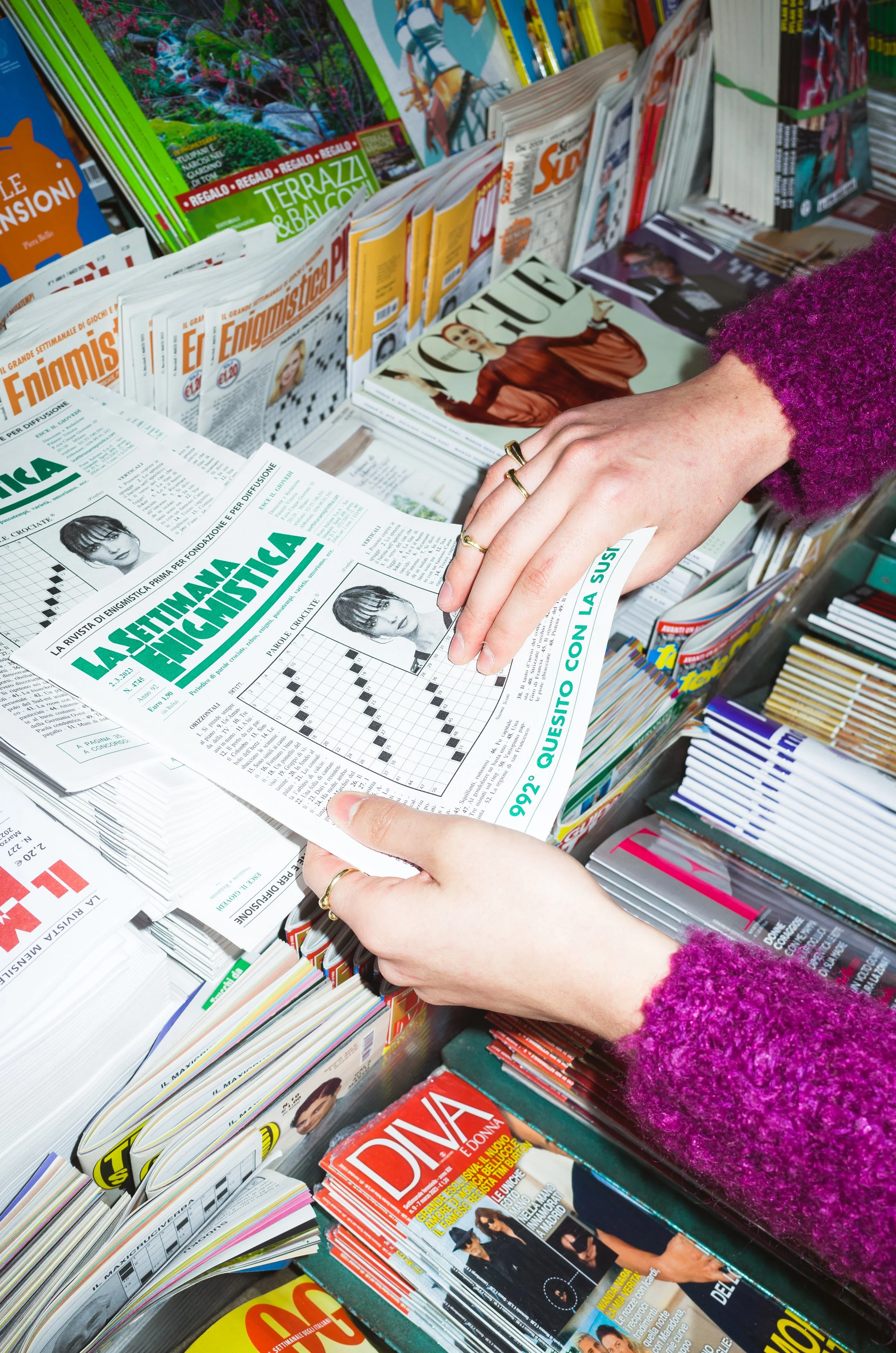 Person browsing magazines and newspapers at a newsstand, holding a crossword puzzle page with a woman's image and text in Italian.