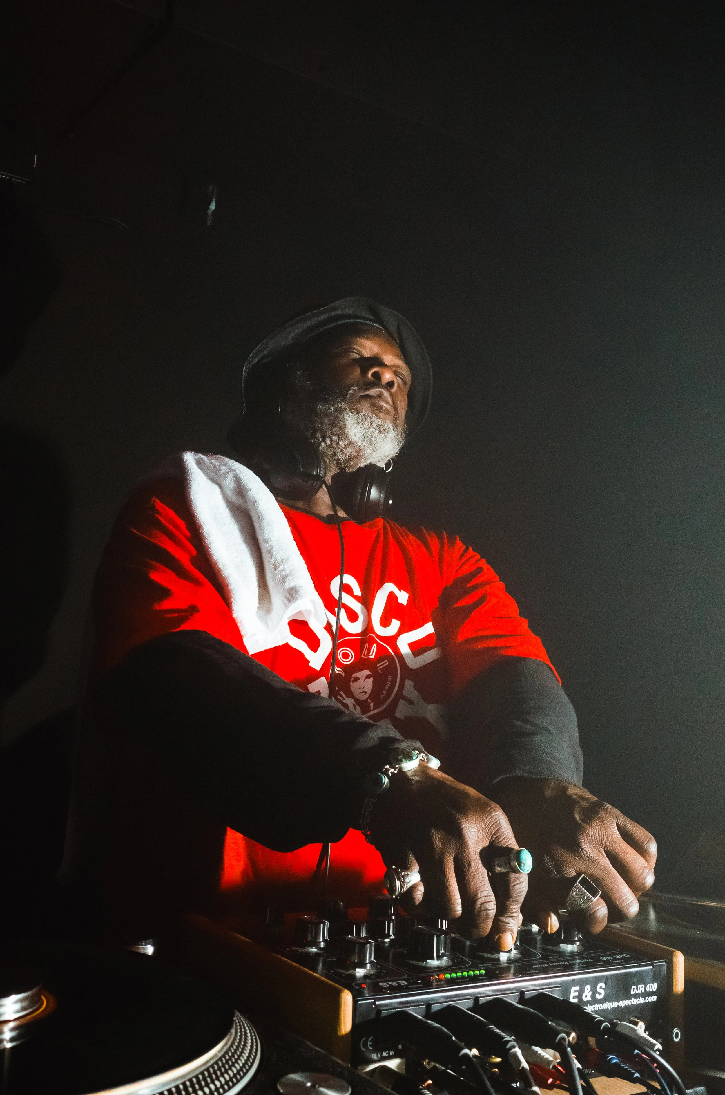 A DJ with headphones and a beard wearing a red and black shirt, standing behind a DJ mixing console in a dark setting.