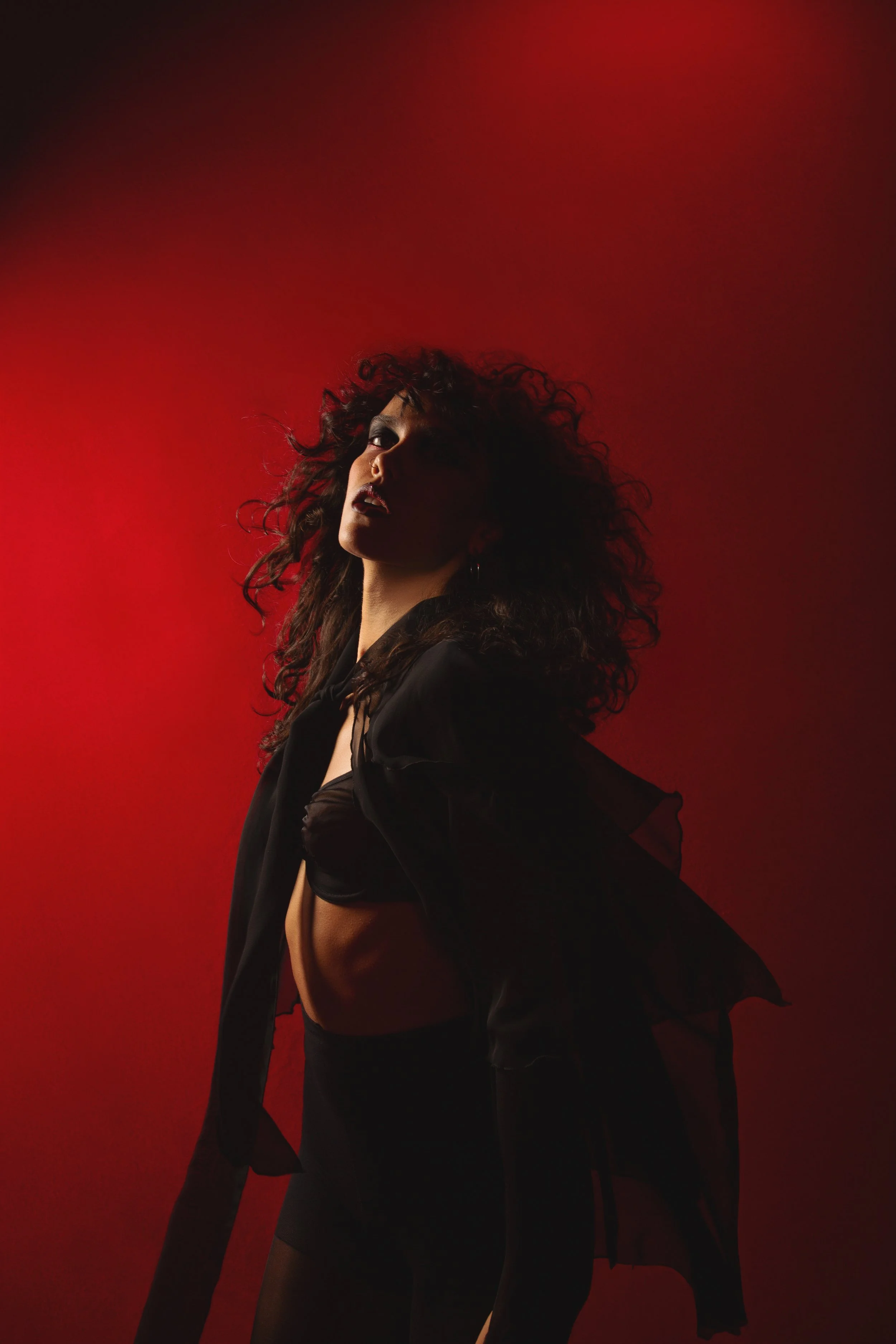 A woman with curly hair posing against a red background, wearing dark clothing and lingerie.