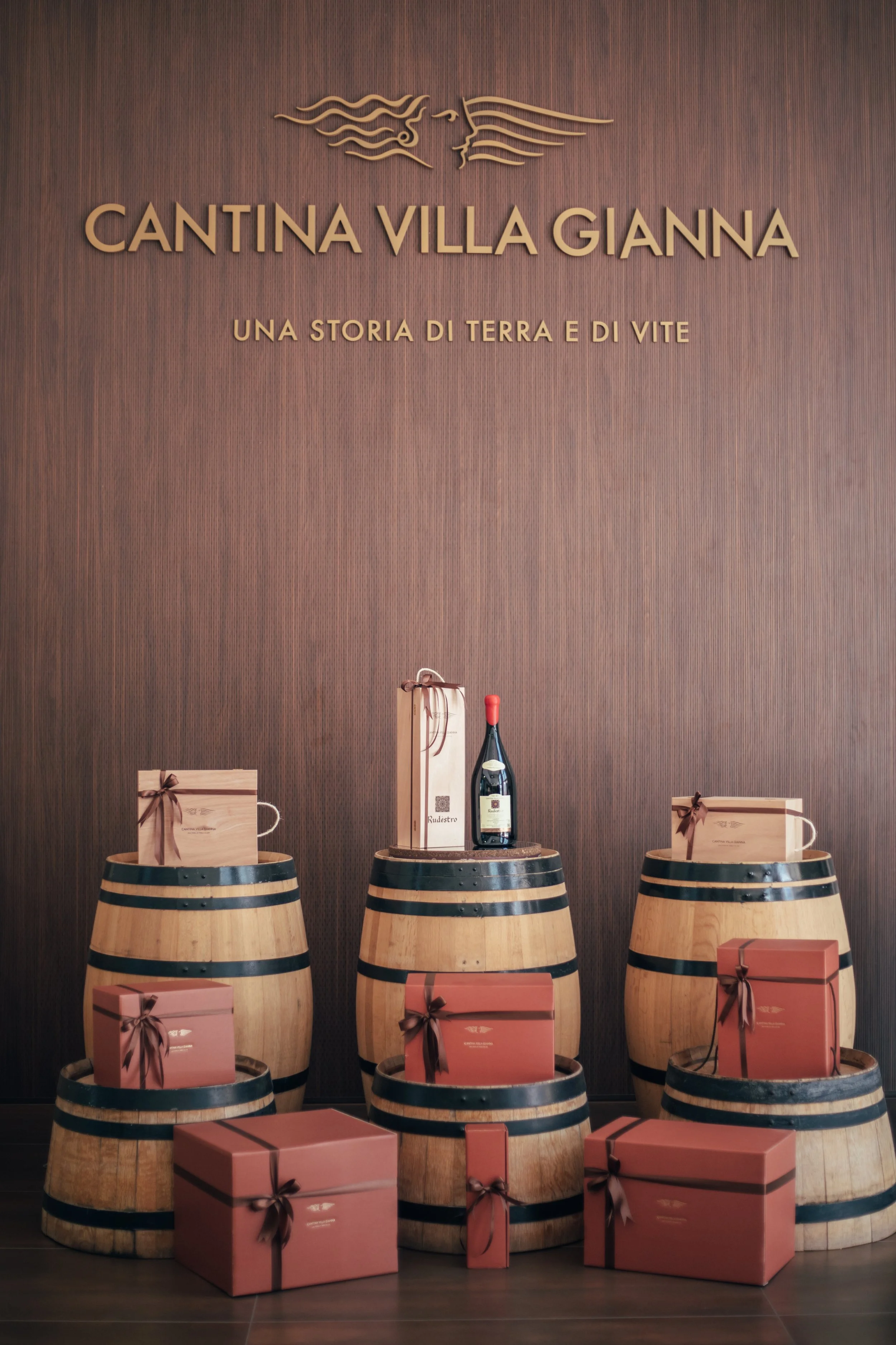 Wooden wine barrels with pink gift boxes and a bottle of wine on top, set against a wood-paneled wall with a sign reading 'Cantina Villa Gianna' and an Italian phrase.