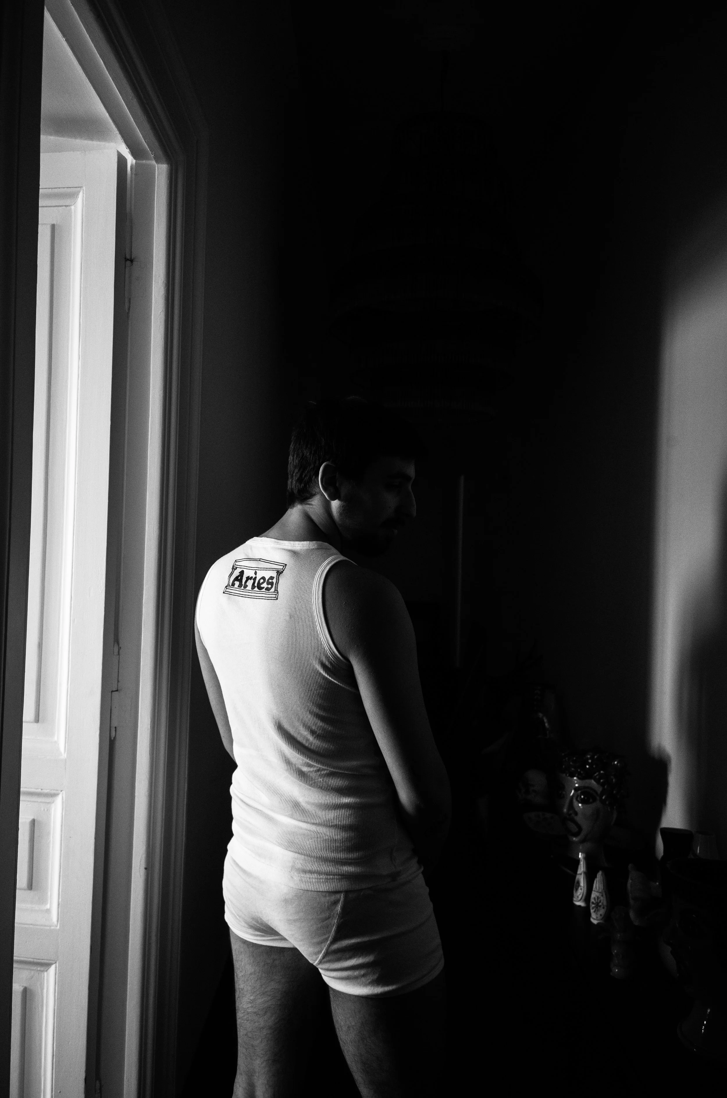 A man in a sleeveless shirt and shorts standing in a dimly lit room near a doorway, with objects and a decorative mask on a table.
