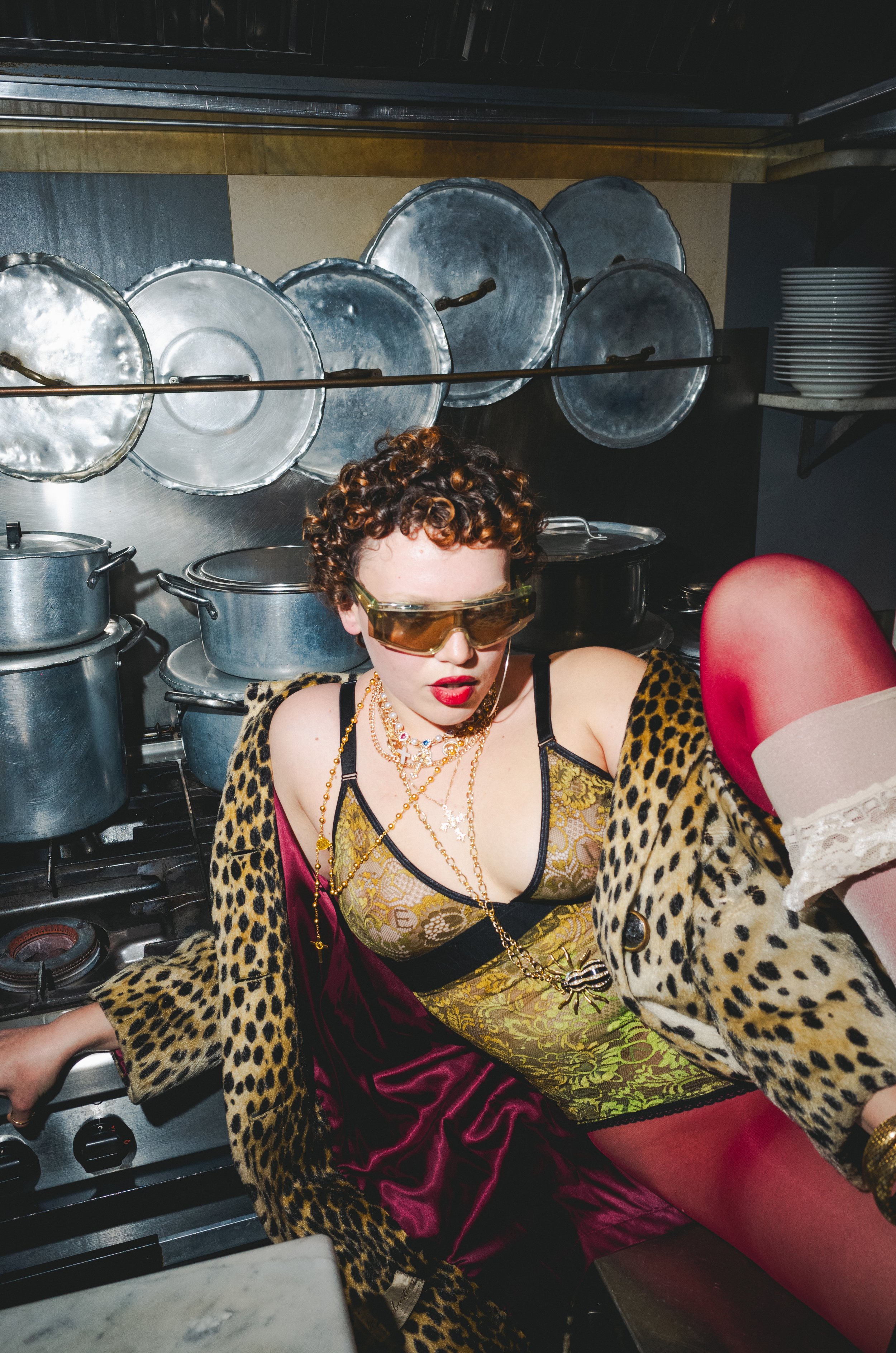 A woman with curly hair and sunglasses in a kitchen, wearing leopard print and vintage style clothing, lounging among pots and pans.