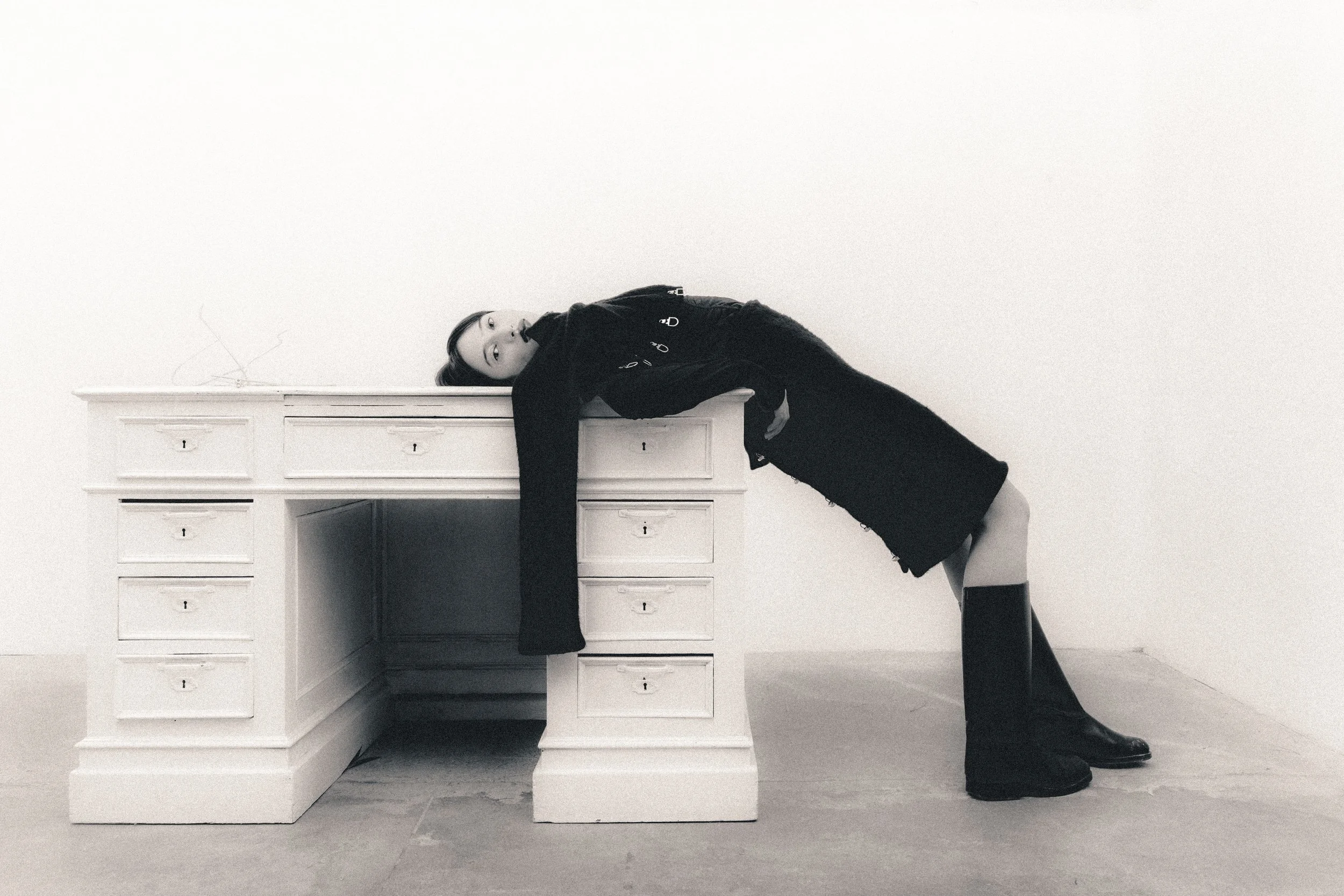 A woman with long hair lying on a desk, with her head and arm hanging off the edge, in a minimalist room with a plain white wall.