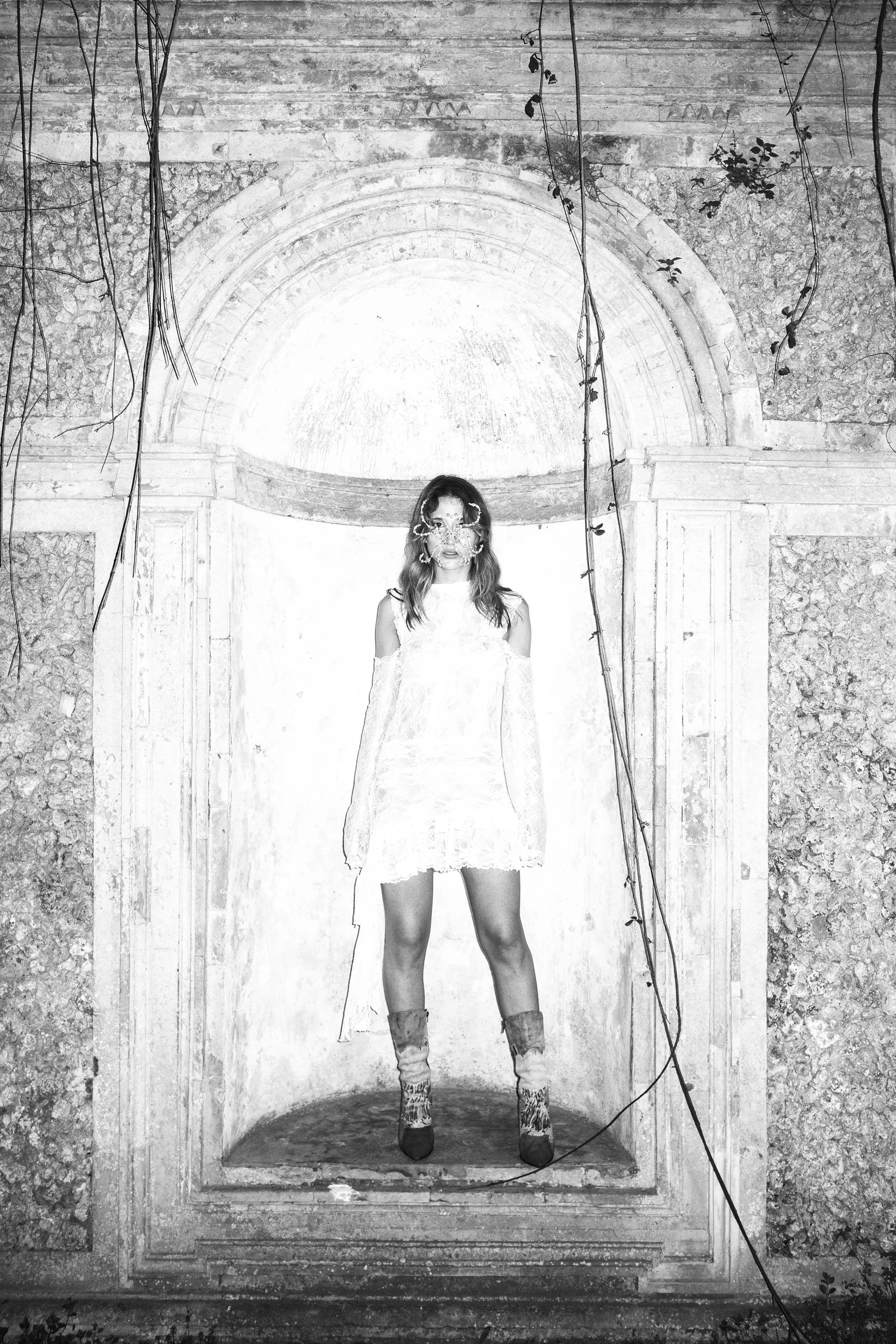 A woman standing in a decorative archway with textured walls and hanging vines, wearing a lace dress and high-heeled boots.