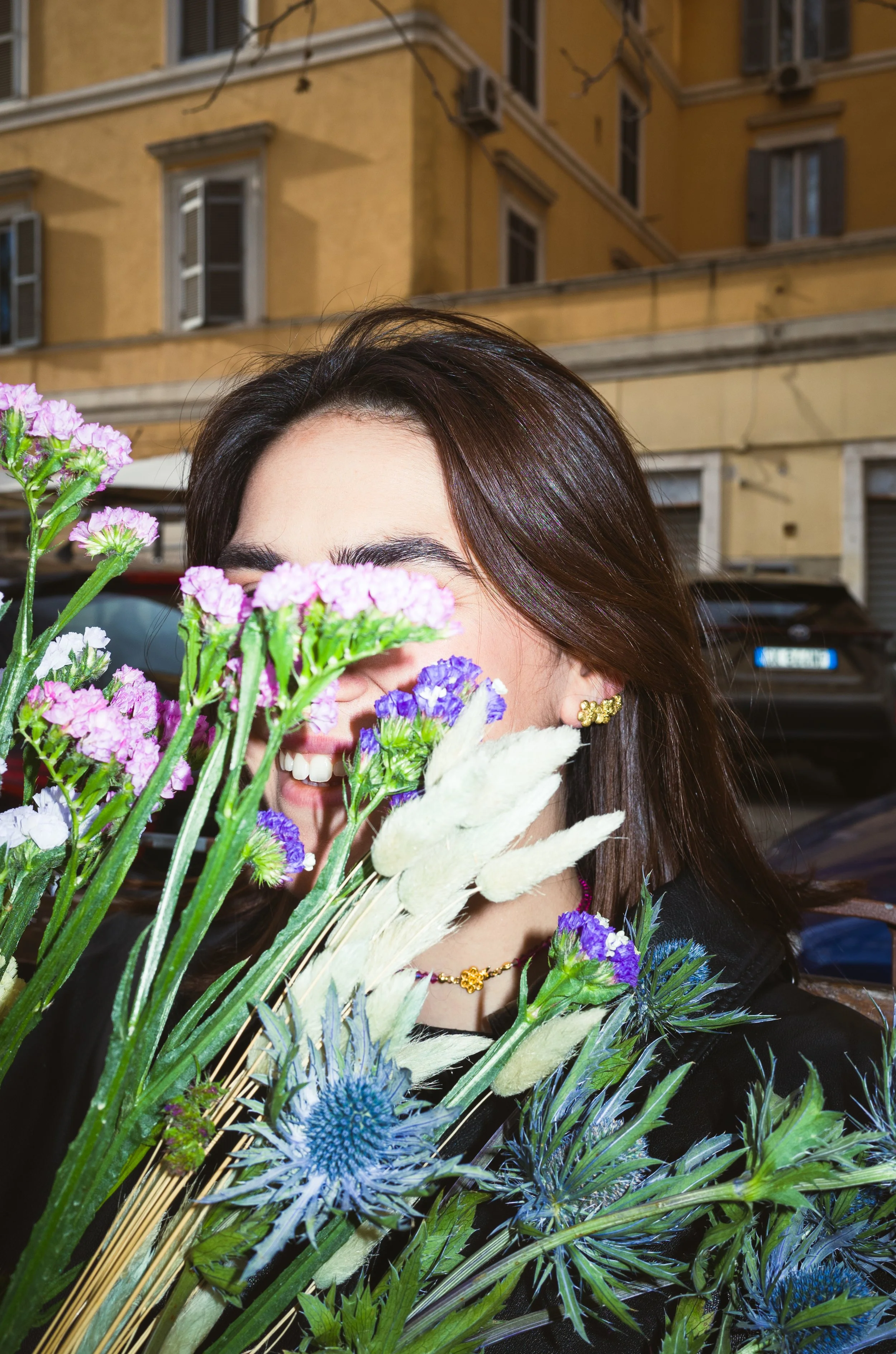A woman with dark hair smiling behind a bouquet of colorful flowers, including purple, blue, and white blossoms, in an urban setting with yellow buildings and parked cars in the background.
