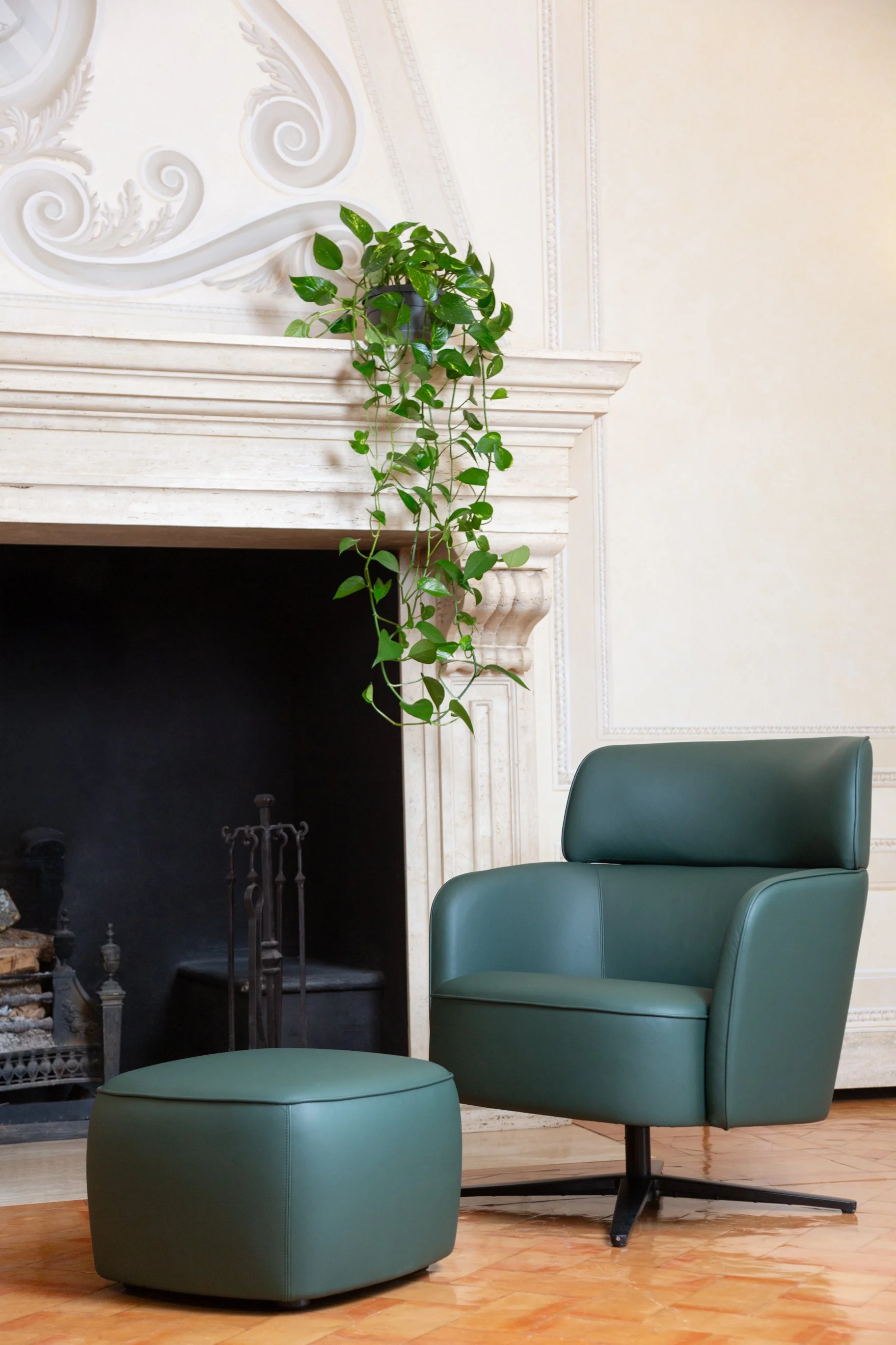 Green armchair and matching ottoman in front of a white fireplace, with a hanging plant on the mantel, cream-colored walls and wooden flooring.