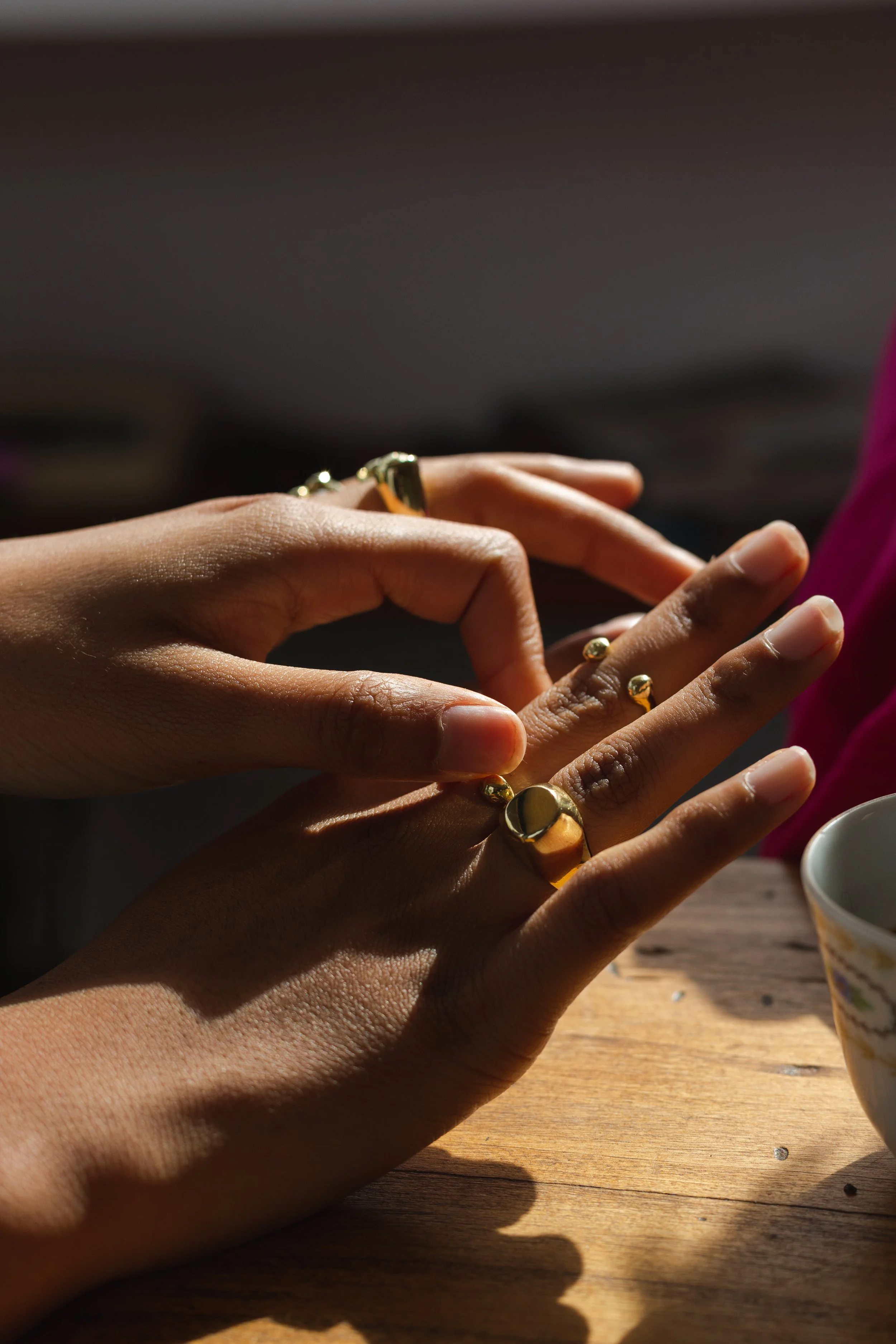 Close-up of a person's hands adjusting gold jewelry, with a wooden table and a ceramic cup nearby, sunlight casting shadows.