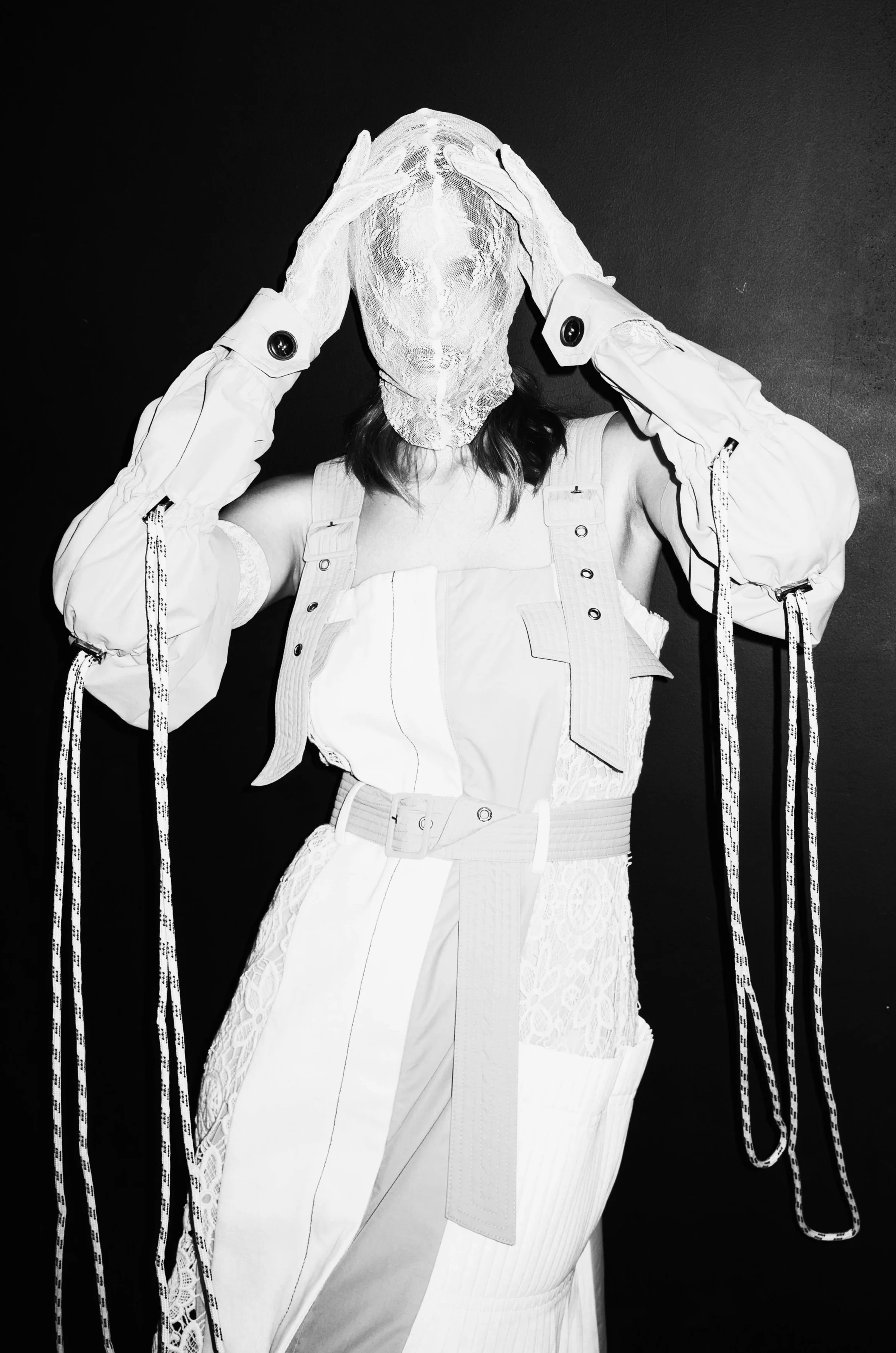 A person wearing a layered white outfit, with a face obscured by a lace mask, holding their head with both hands, set against a dark background.