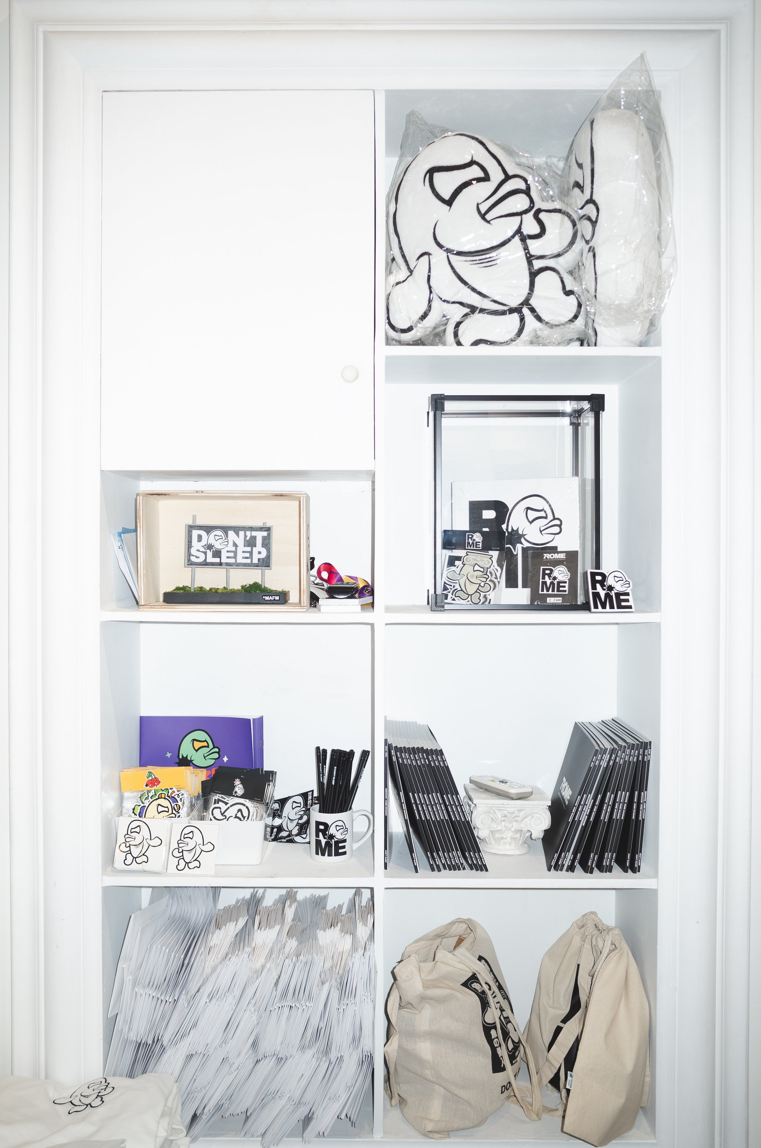 White shelving unit filled with black and white cartoon duck-themed merchandise, stationery, tote bags, and artwork, featuring items like notebooks, posters, and plush toys.