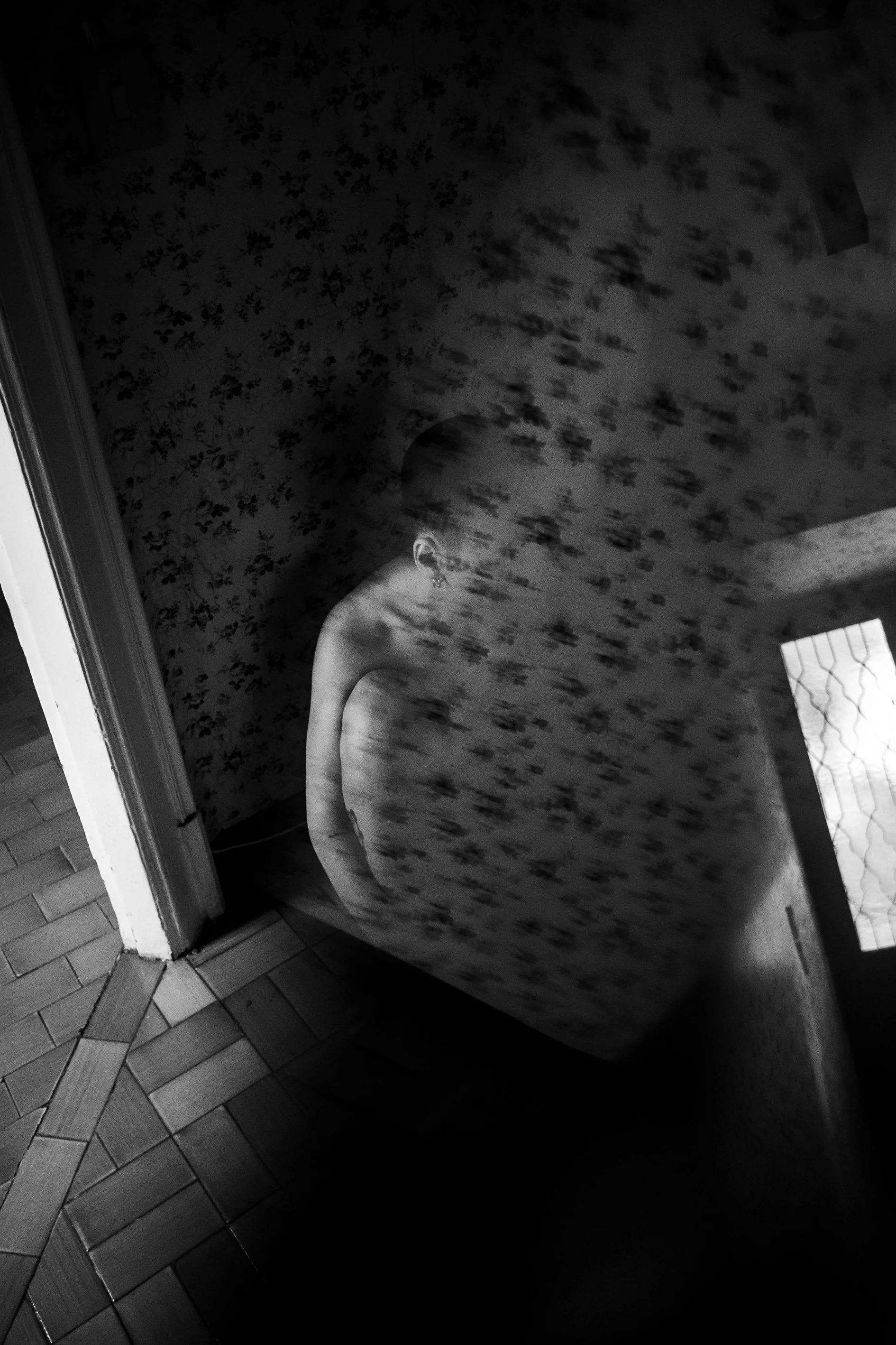Black and white photo of a person with short hair, standing in front of a patterned wall, reflected in a mirror, with a window showing blinds on the side.