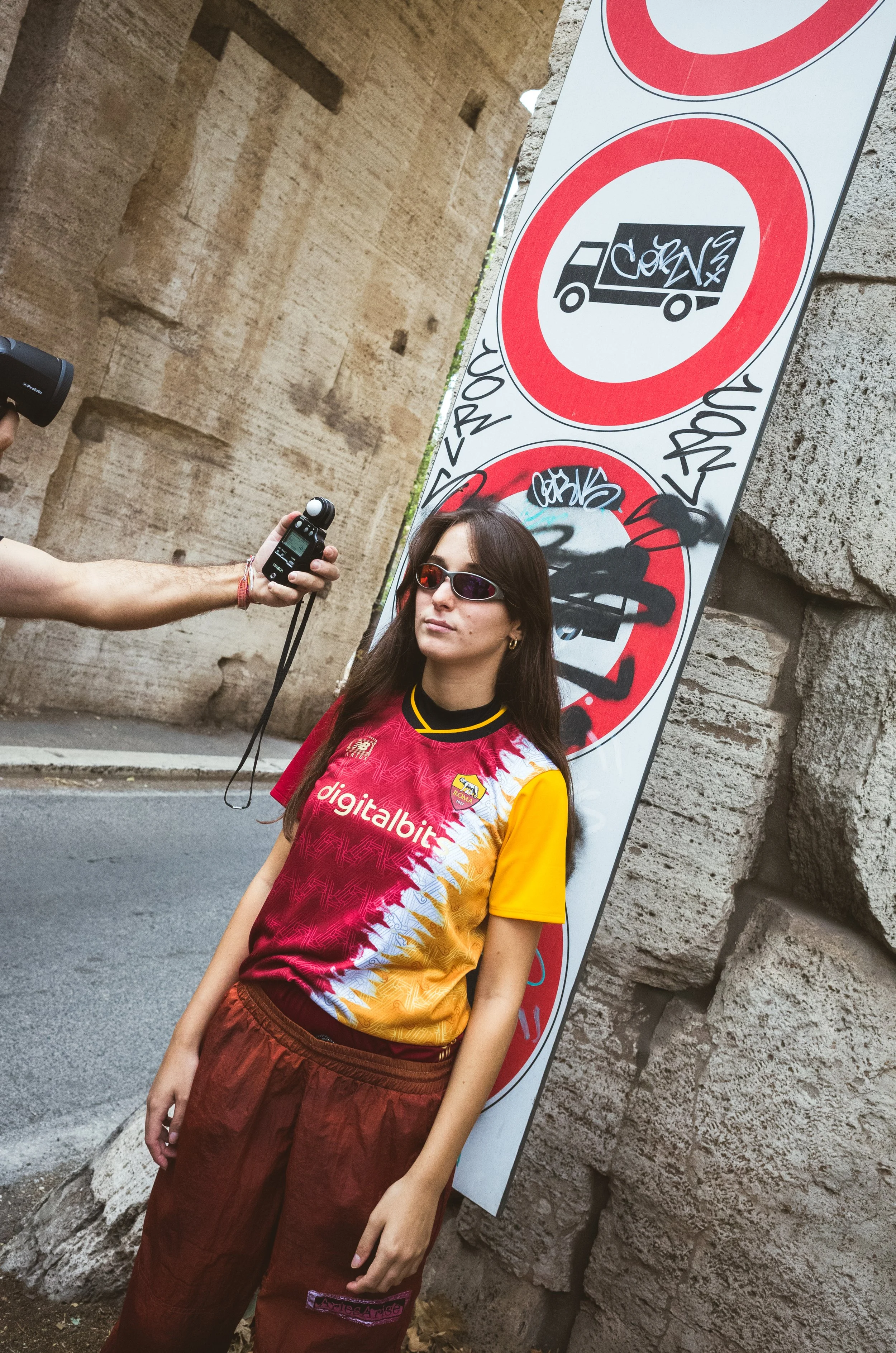 A woman in a colorful sports jersey and red pants is standing next to a graffiti-covered sign on a stone wall, while a person holds a recording device toward her. The sign indicates restrictions on trucks, graffiti, and other activities.