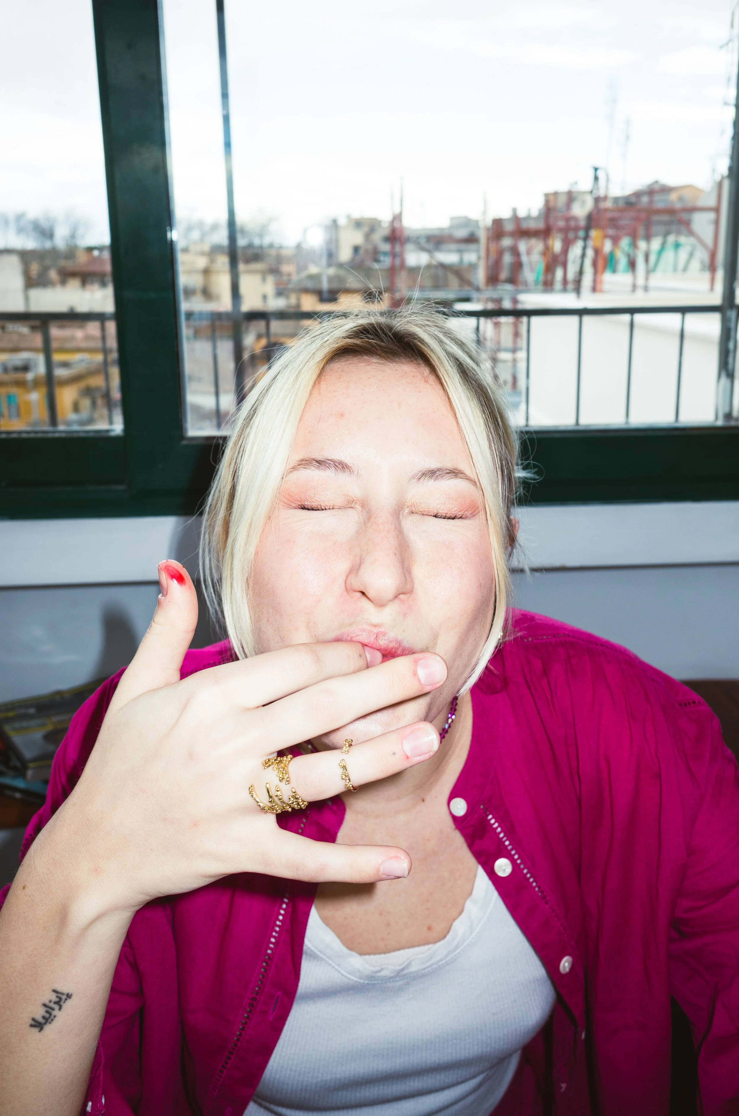 A woman with blonde hair wearing a red shirt with white underneath, with her eyes closed, covering her mouth with her hand, inside a room with large windows showing rooftops and a cloudy sky outside.