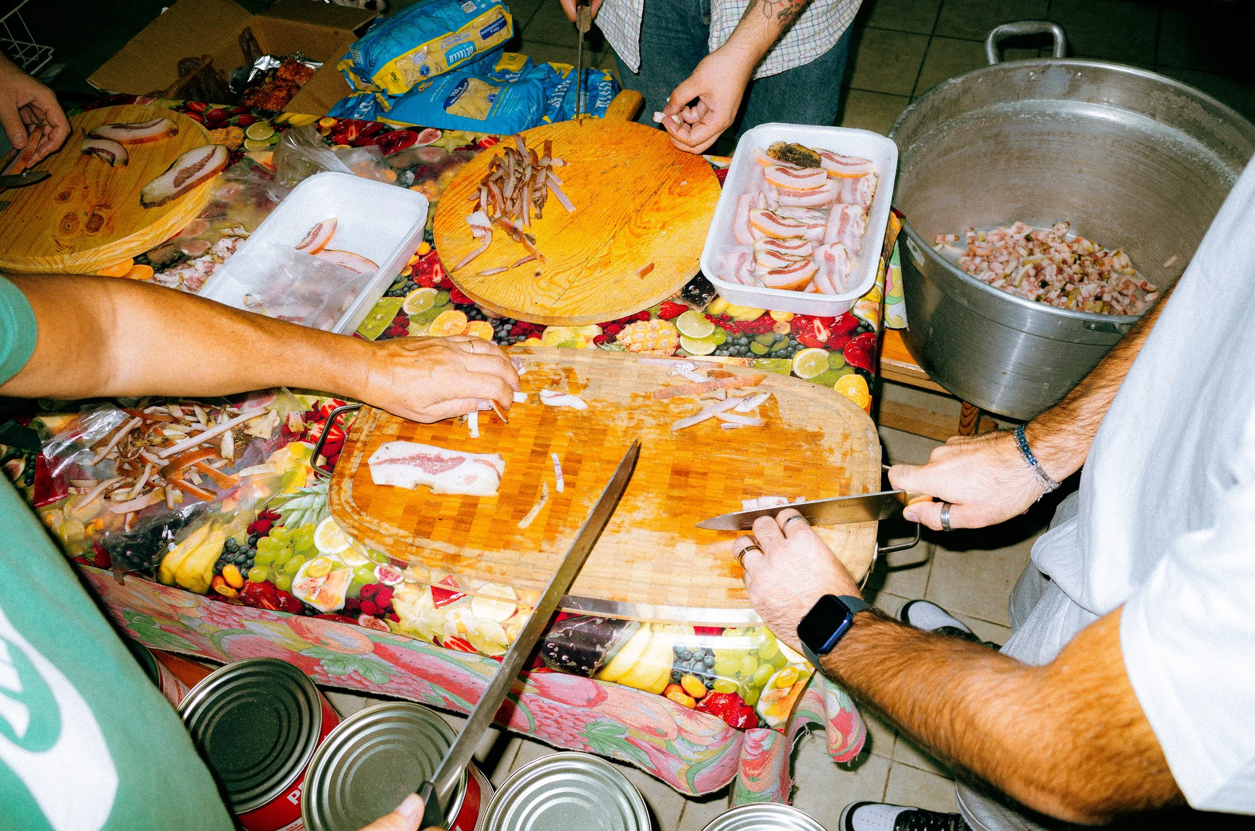 People preparing bacon and mushrooms on a kitchen table with a colorful tablecloth, using knives and cutting boards, with canned food and packaging nearby.