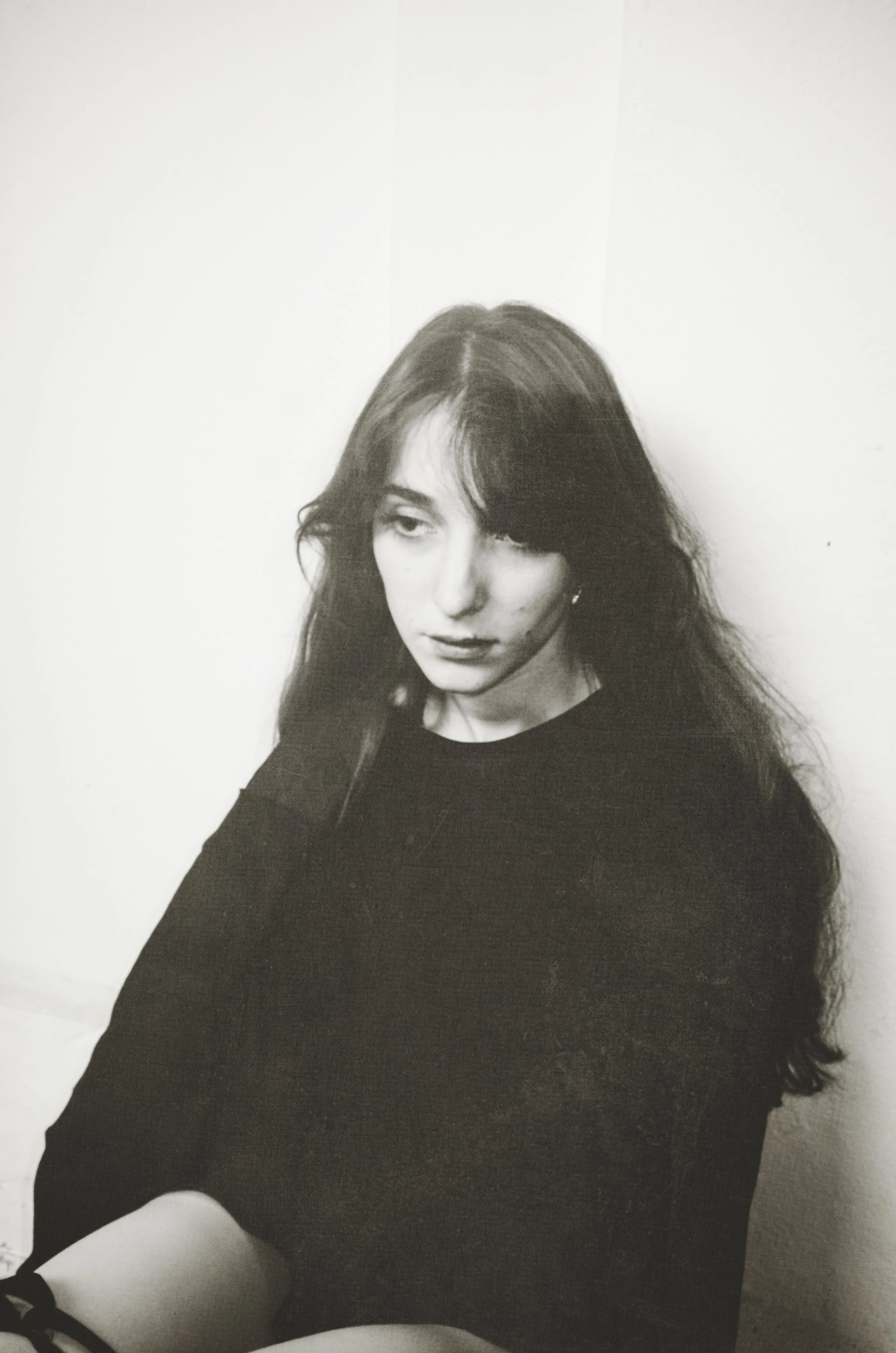 Black and white photograph of a young woman with long, dark hair, sitting against a plain white wall. She is looking off to the side with a thoughtful expression, wearing a dark sweater, with her right hand resting on her neck.