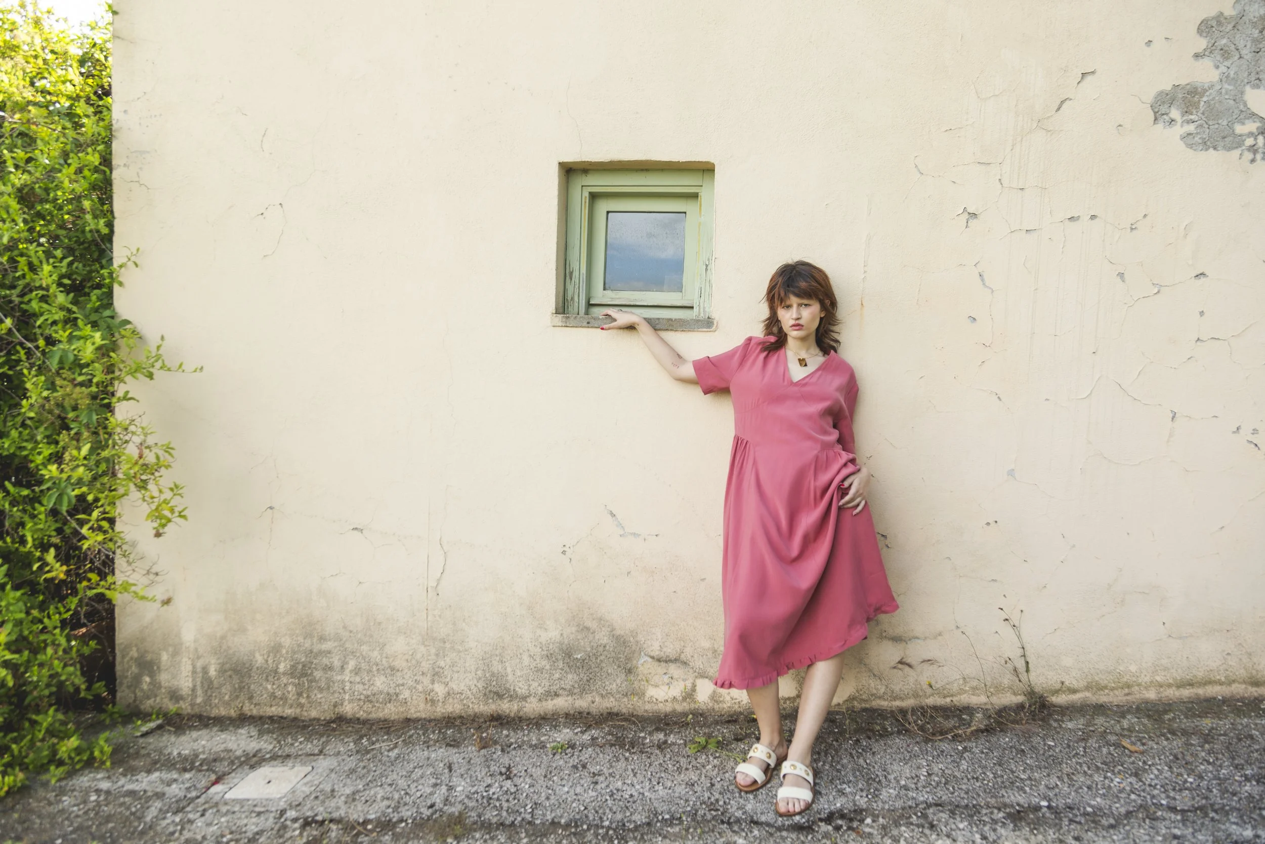 A woman in a pink dress and white sandals leaning against a textured, off-white wall with a small green window and some greenery to the side.