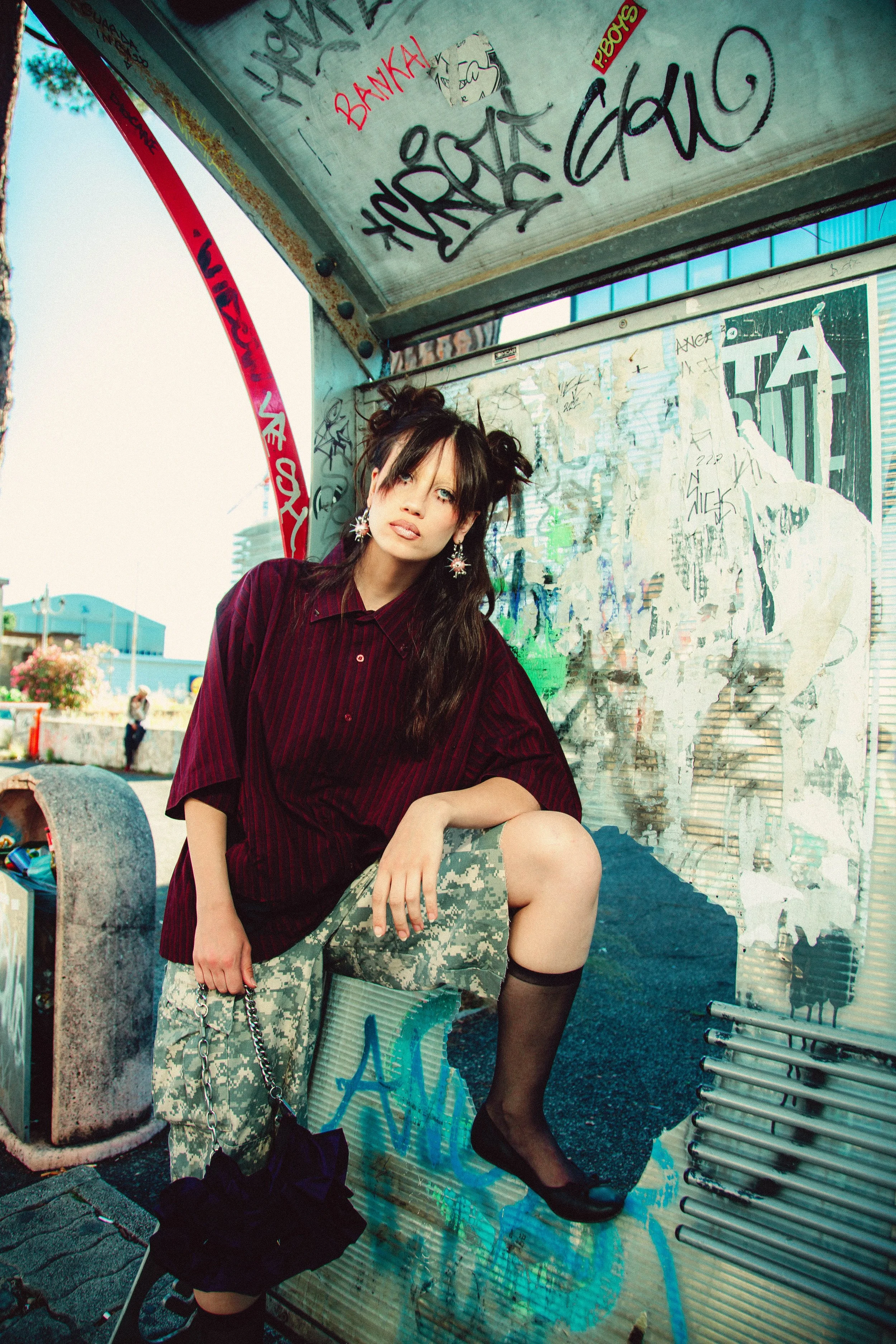 A young woman with dark hair styled in two buns, wearing a red and black striped shirt, camouflage shorts, and black knee-high socks, posing in a graffiti-covered bus stop or shelter.