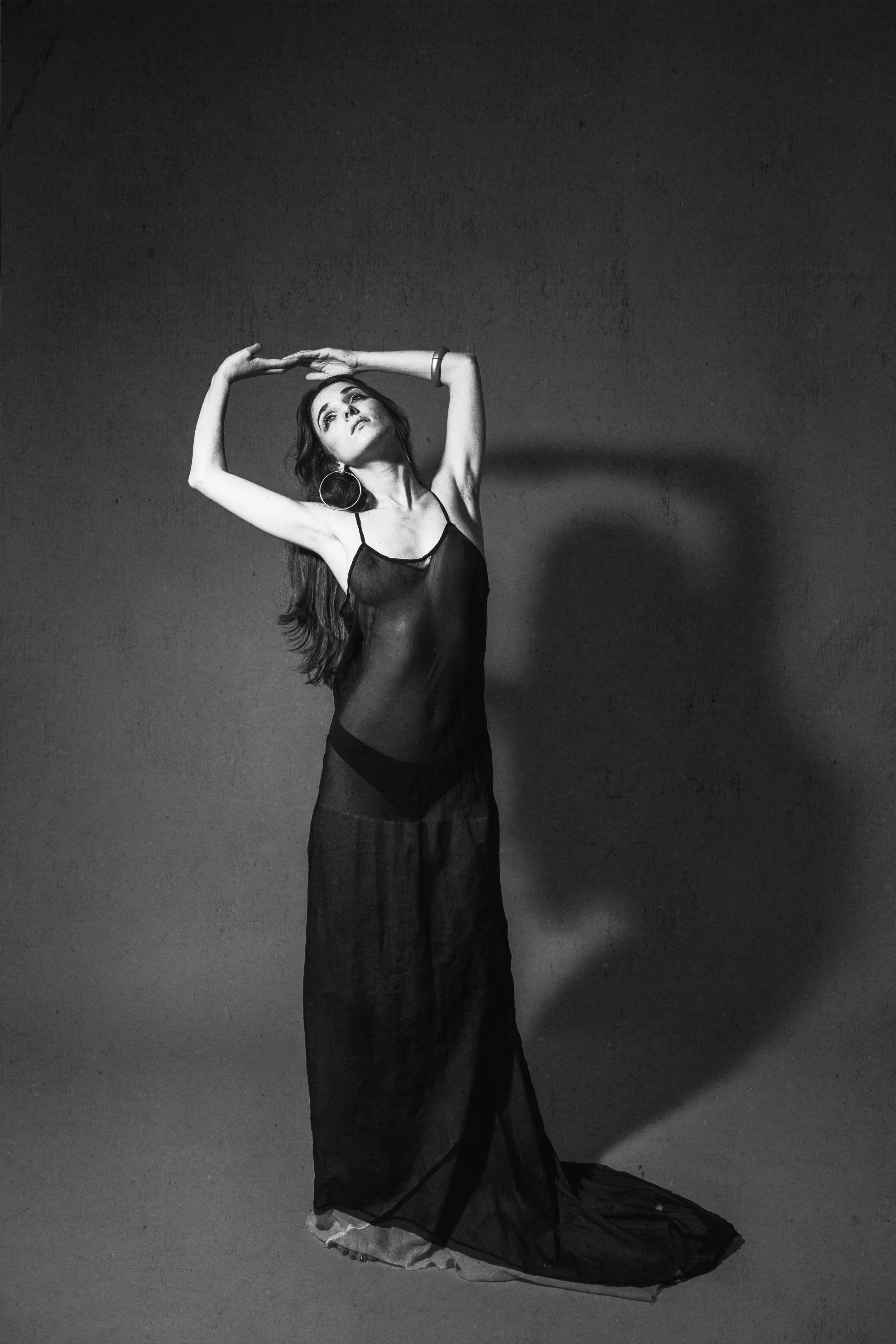 A woman in a sheer black dress posing with arms raised above her head, casting a shadow on the wall behind her in a studio setting.