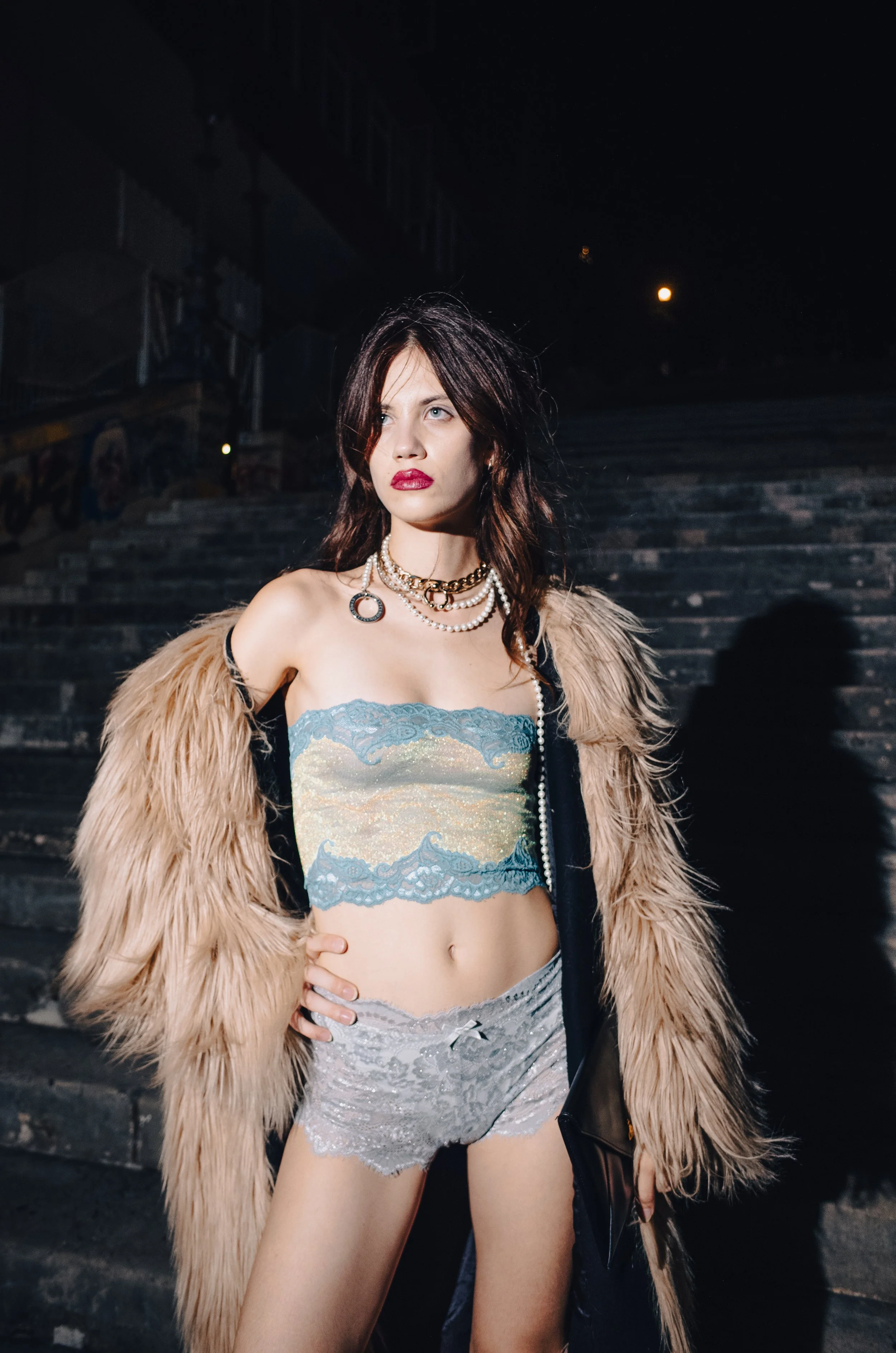 Fashionably dressed woman with dark hair, wearing jewelry, a faux fur coat, and shorts, standing outdoors at night on steps.