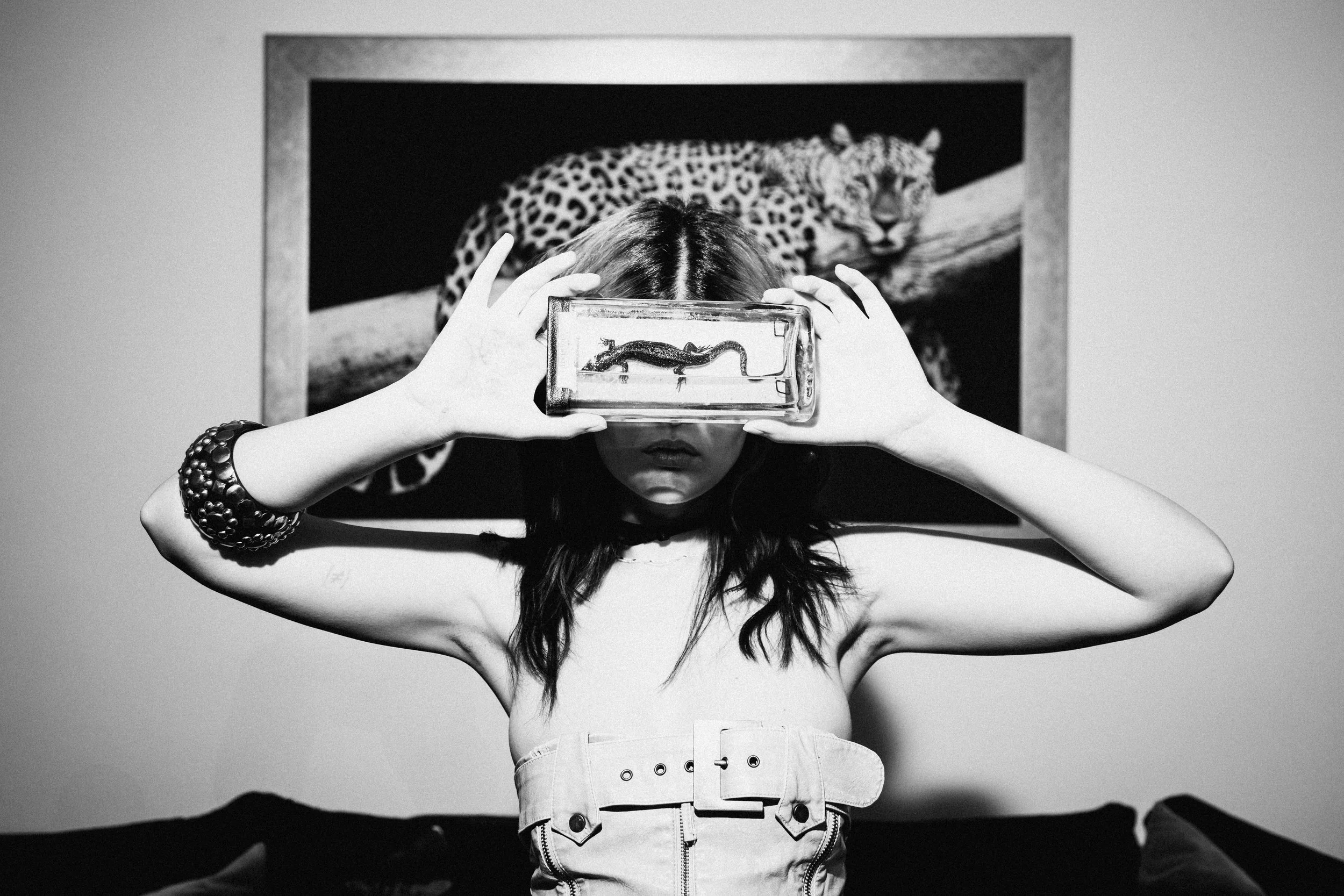 A woman holds a glass container with a small lizard against her face, obscuring her eyes, in front of a photograph of a leopard in the background.