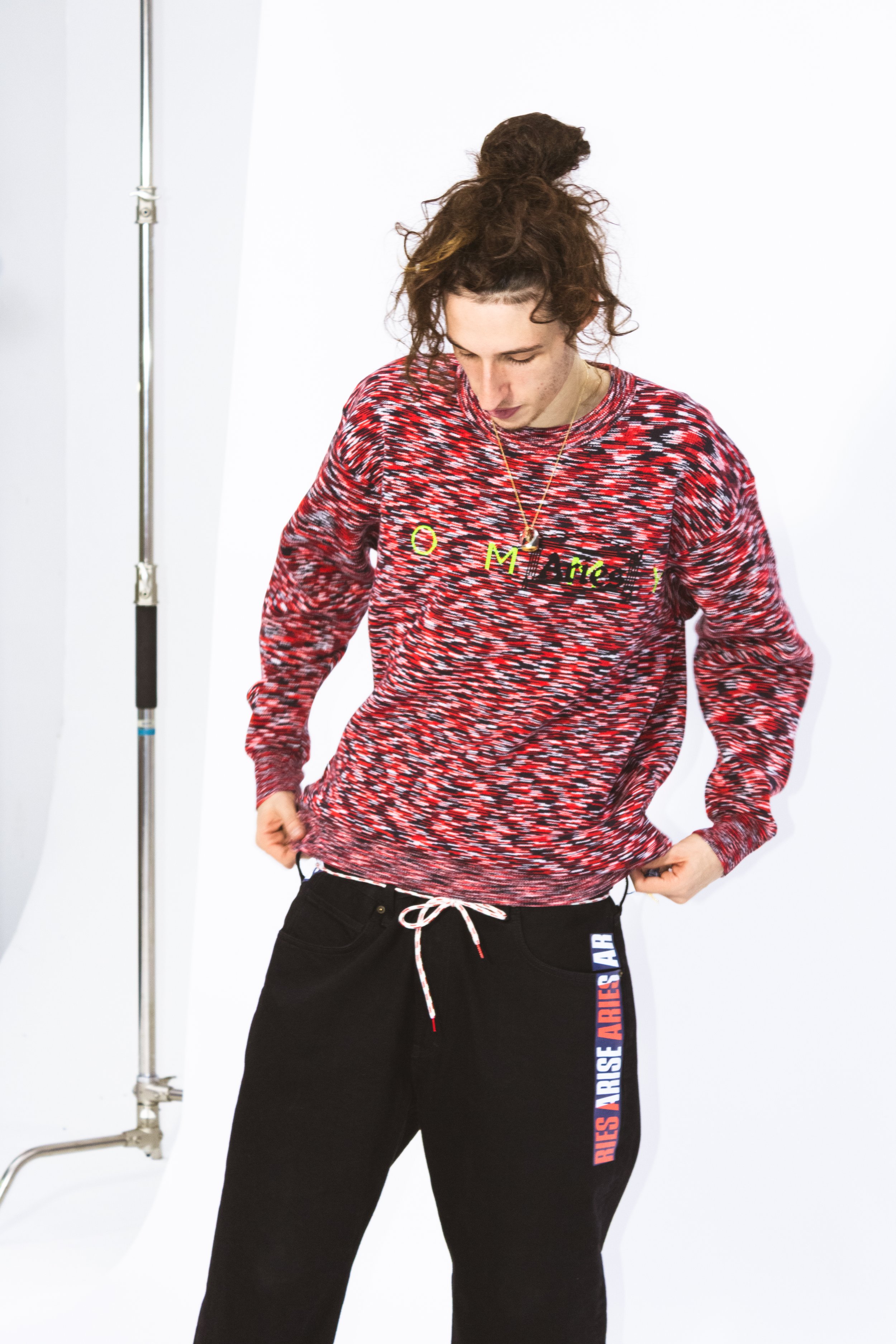 A young woman with curly brown hair tied up in a messy bun wearing a patterned red, black, and white sweatshirt with the words 'O Come' on it, and black sweatpants with the words 'DIES ARISE' printed on the side, standing against a white background.