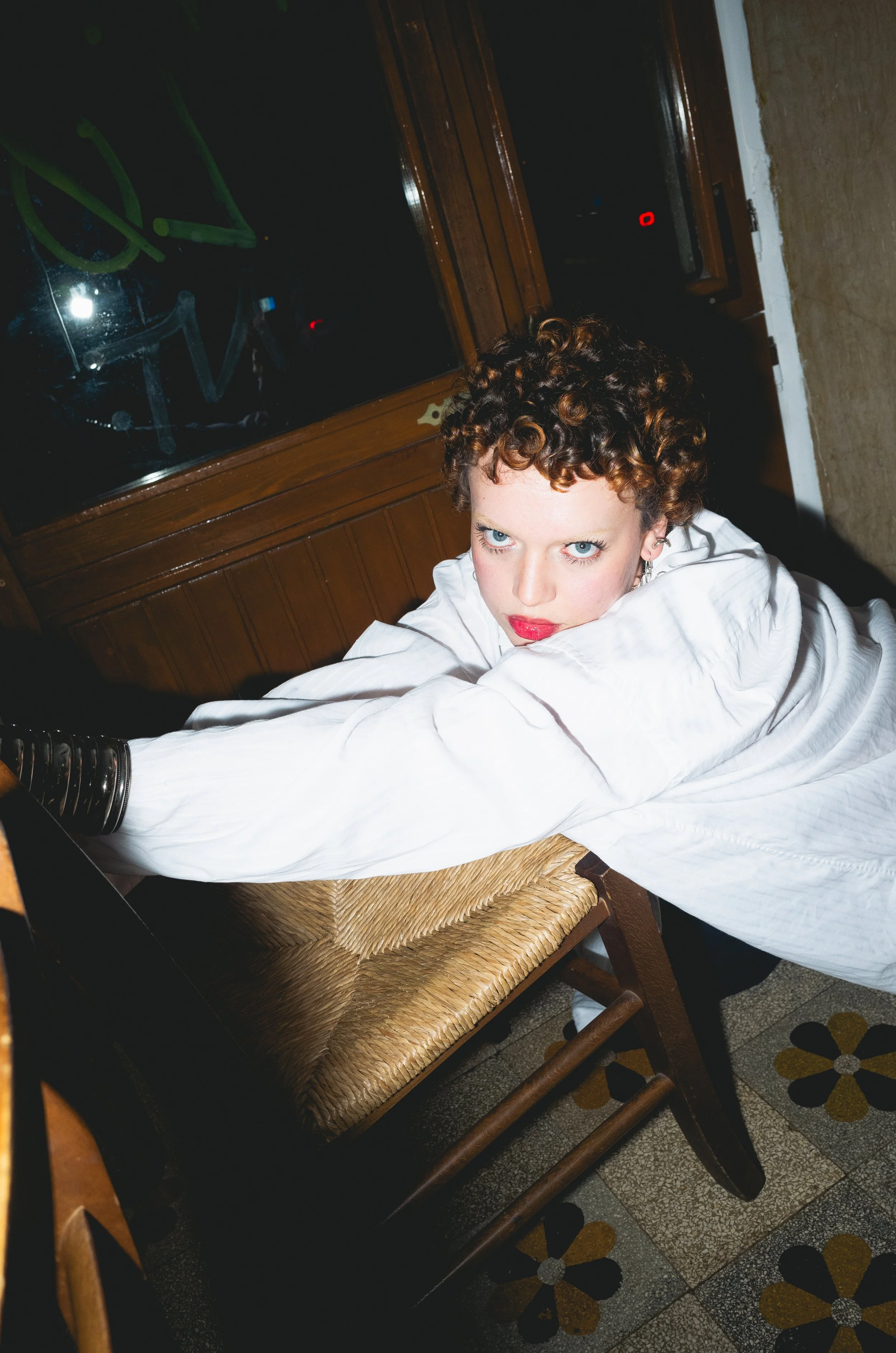 A woman with short curly hair, wearing a white shirt, gazes intensely at the camera as she leans over a wooden chair in a dimly lit room with patterned tile flooring and a glass window or door in the background.