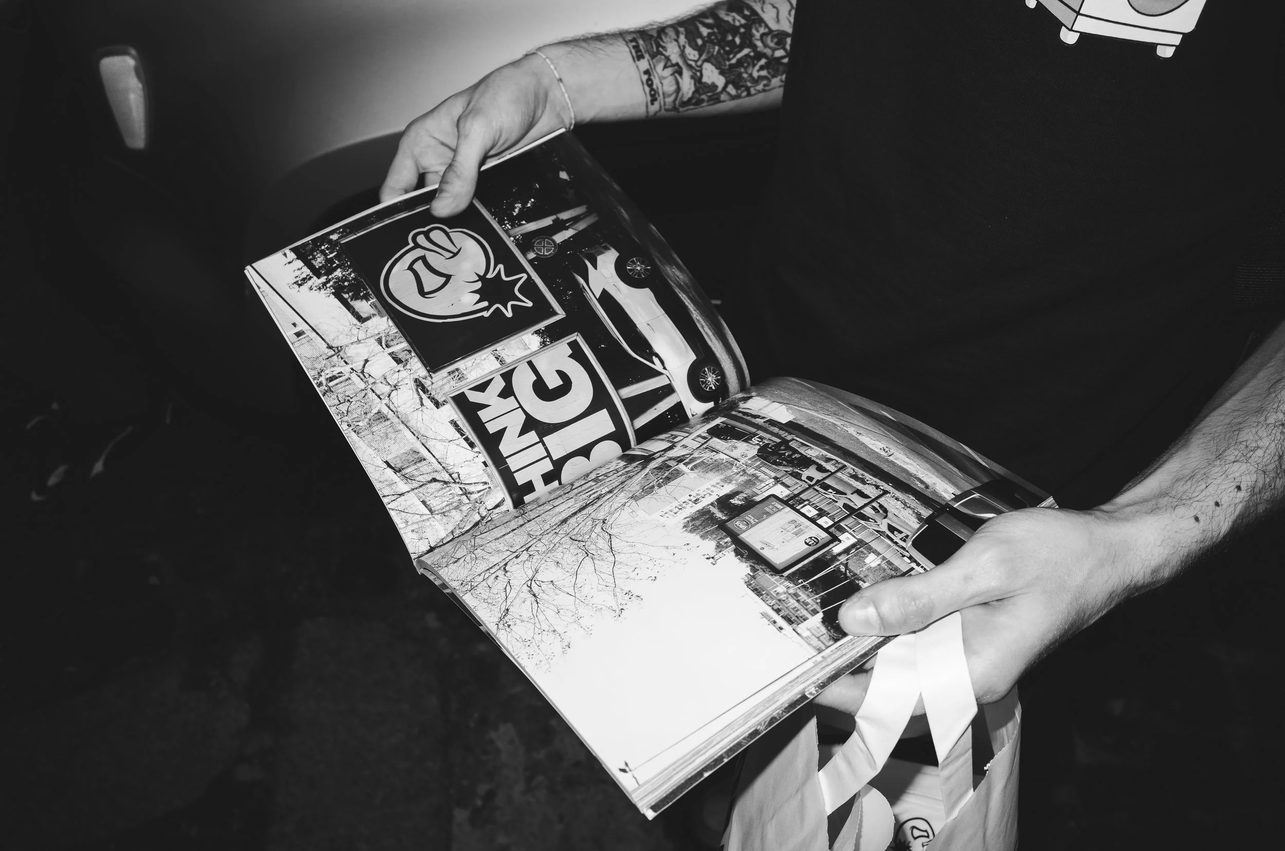 Person holding an open magazine with street scenes, a parked car, and stickers, including one with a fist and bomb icon, in black and white.