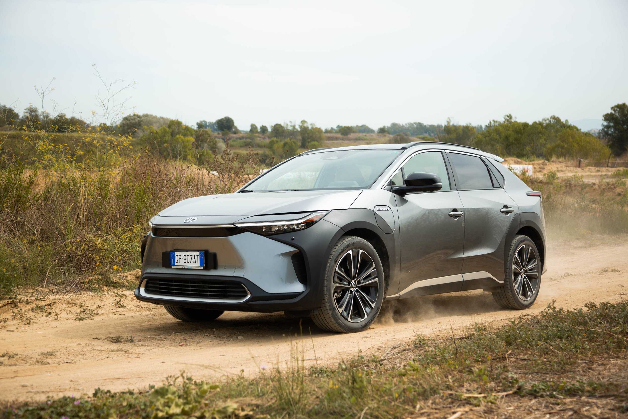 A gray electric SUV driving on a dirt road in a rural area with bushes and trees in the background.