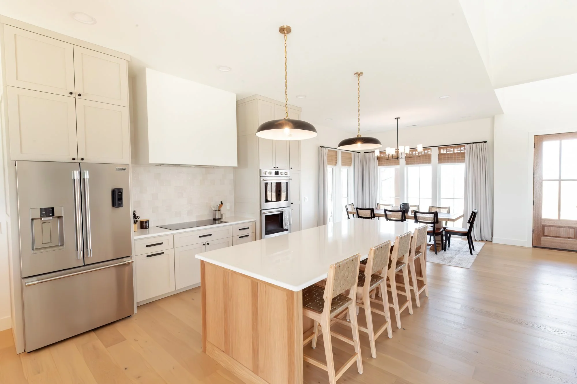 A modern kitchen with a large island, white cabinets, stainless steel refrigerator, and a dining area with a table and chairs, illuminated by pendant lights and natural light from large windows.