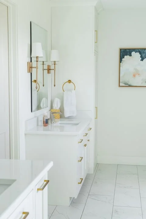 White bathroom vanity with gold hardware, a large mirror, two wall-mounted light fixtures, a gold towel ring with a white towel, a soap dispenser, tissue box, and other toiletries, with white cabinetry and a framed abstract painting on the wall.
