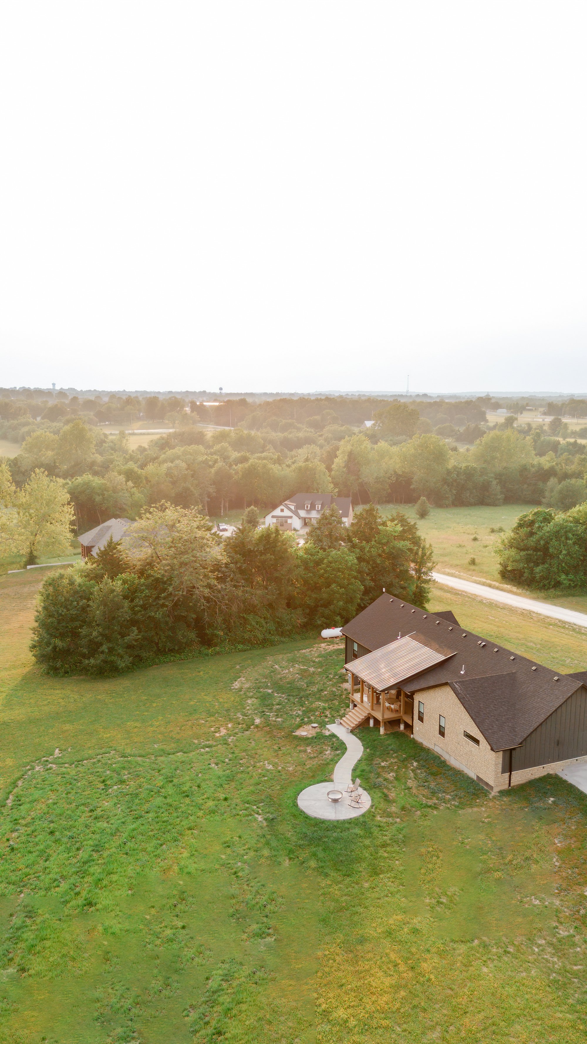 A bird's eye view of a suburban neighborhood with a house, a paved pathway, and a backyard with a fire pit and chairs, surrounded by green trees and grass.