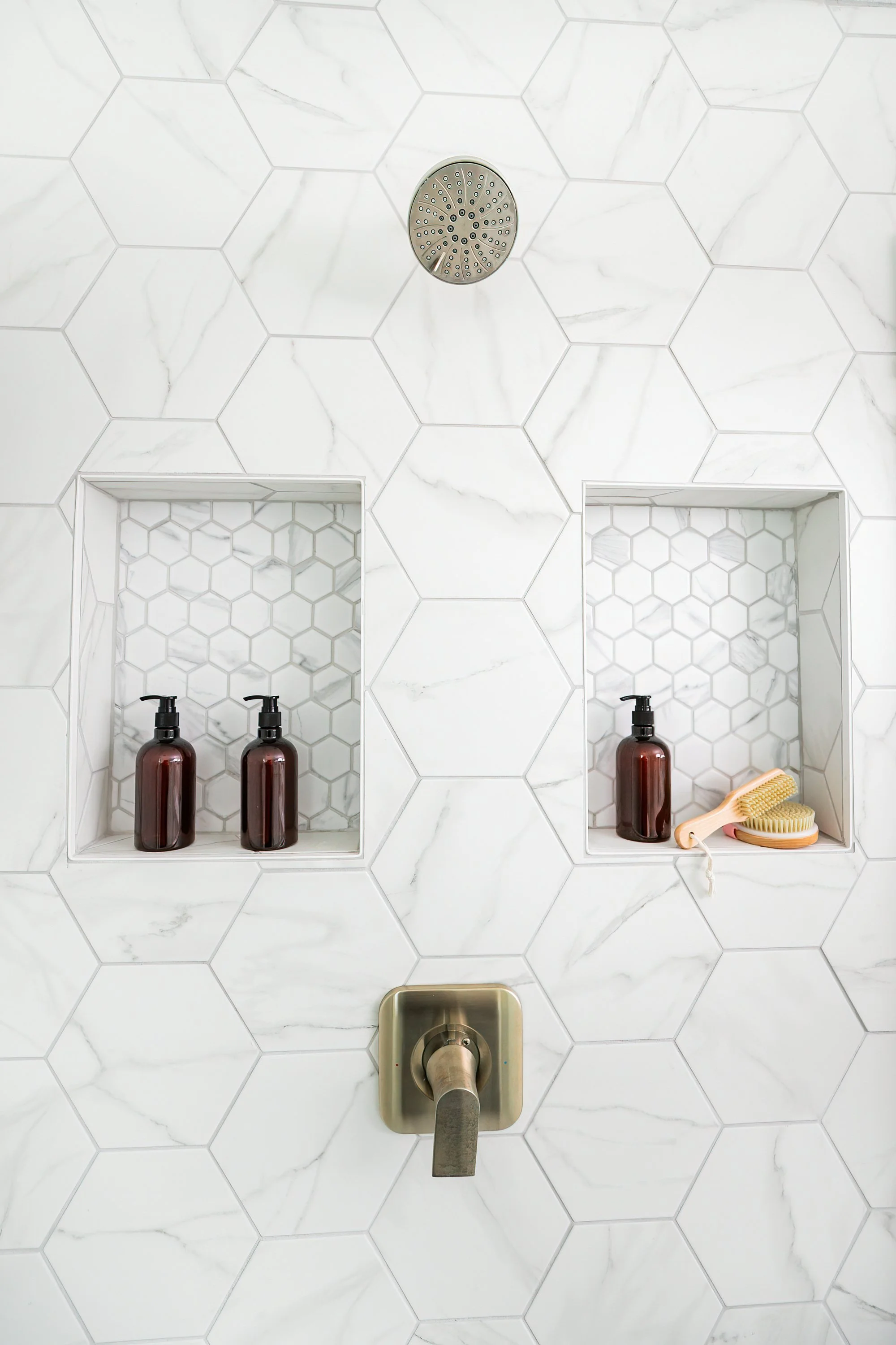 A shower wall with white hexagonal tiles and two built-in recessed shelves. The left shelf contains two dark-colored bottles with pump dispensers. The right shelf has a dark bottle, a yellow bath brush, and a light-colored scrubbing brush. A shower faucet with a silver handle is at the bottom of the image.