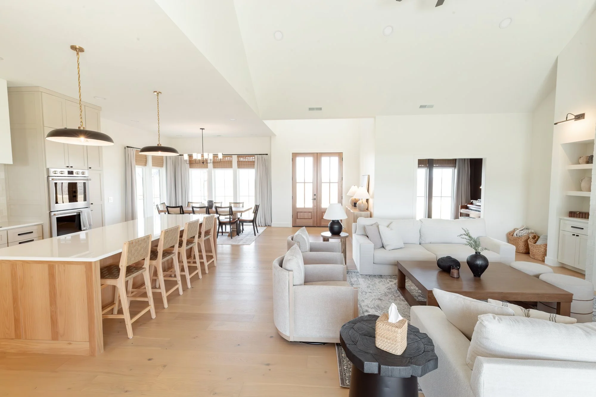 Bright open-concept living room and dining area with white walls, hardwood floors, and natural light. Features include a white L-shaped sectional sofa, a rustic wooden coffee table, black decorative vases, and a side table with lamps. The dining area has a large table with chairs, and there are multiple windows with curtains and blinds. The kitchen area with pendant lights and built-in appliances is partially visible.