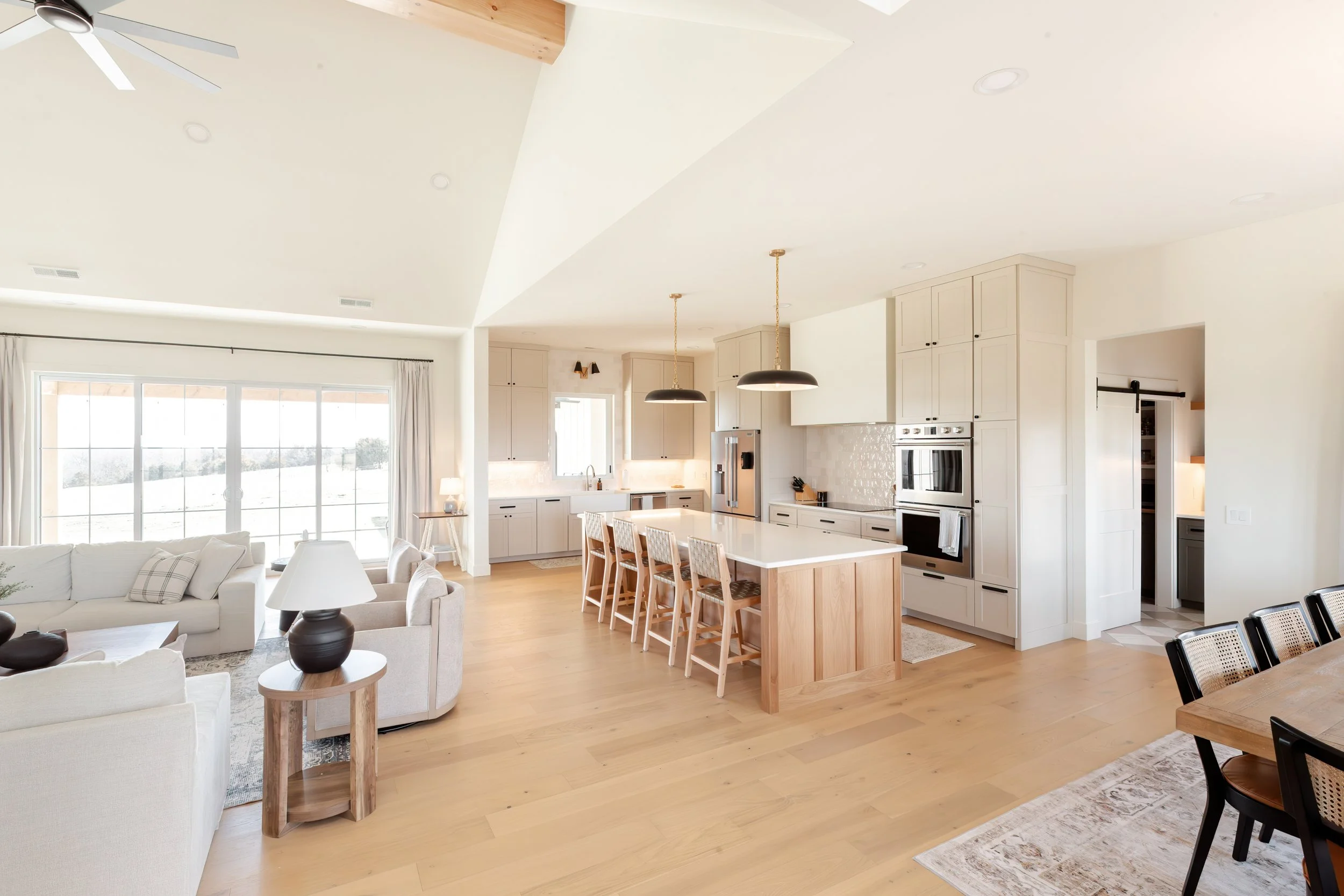 Open-plan living room and kitchen with white cabinets, a kitchen island with wooden barstools, large windows, and modern lighting fixtures.