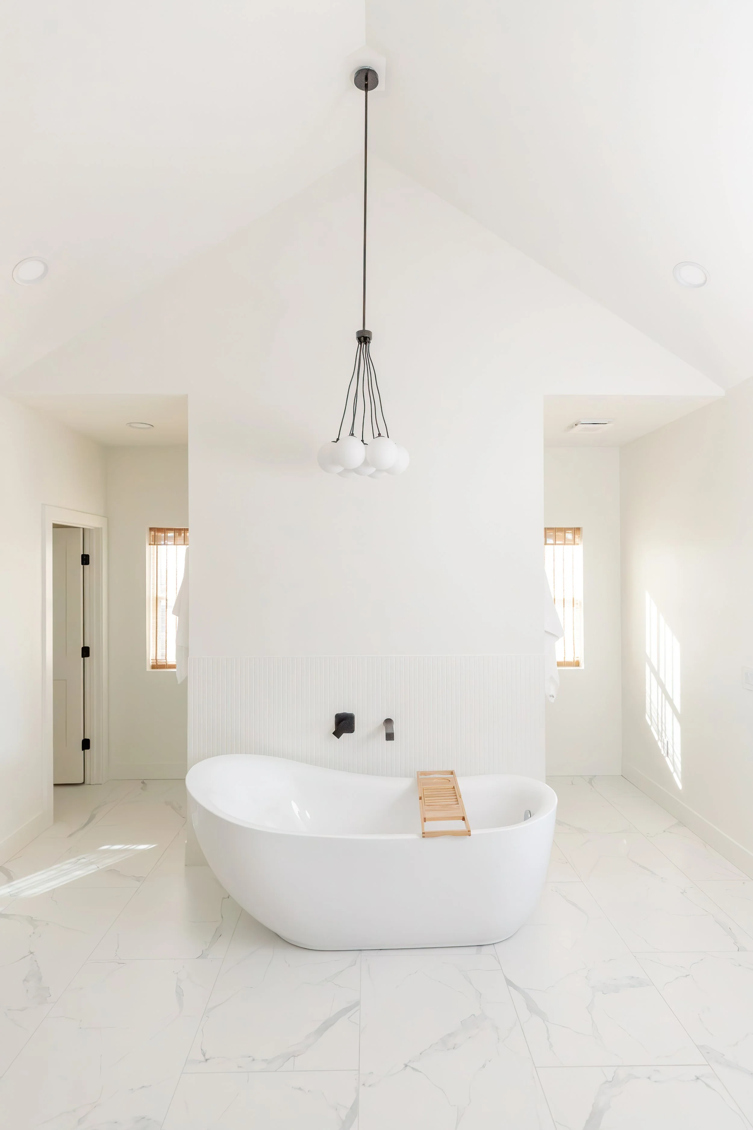A white freestanding bathtub with a wooden bath tray in a bright, minimalist bathroom. The room features white walls, marble tile flooring, two windows with wooden blinds, and a modern black chandelier hanging from the ceiling.