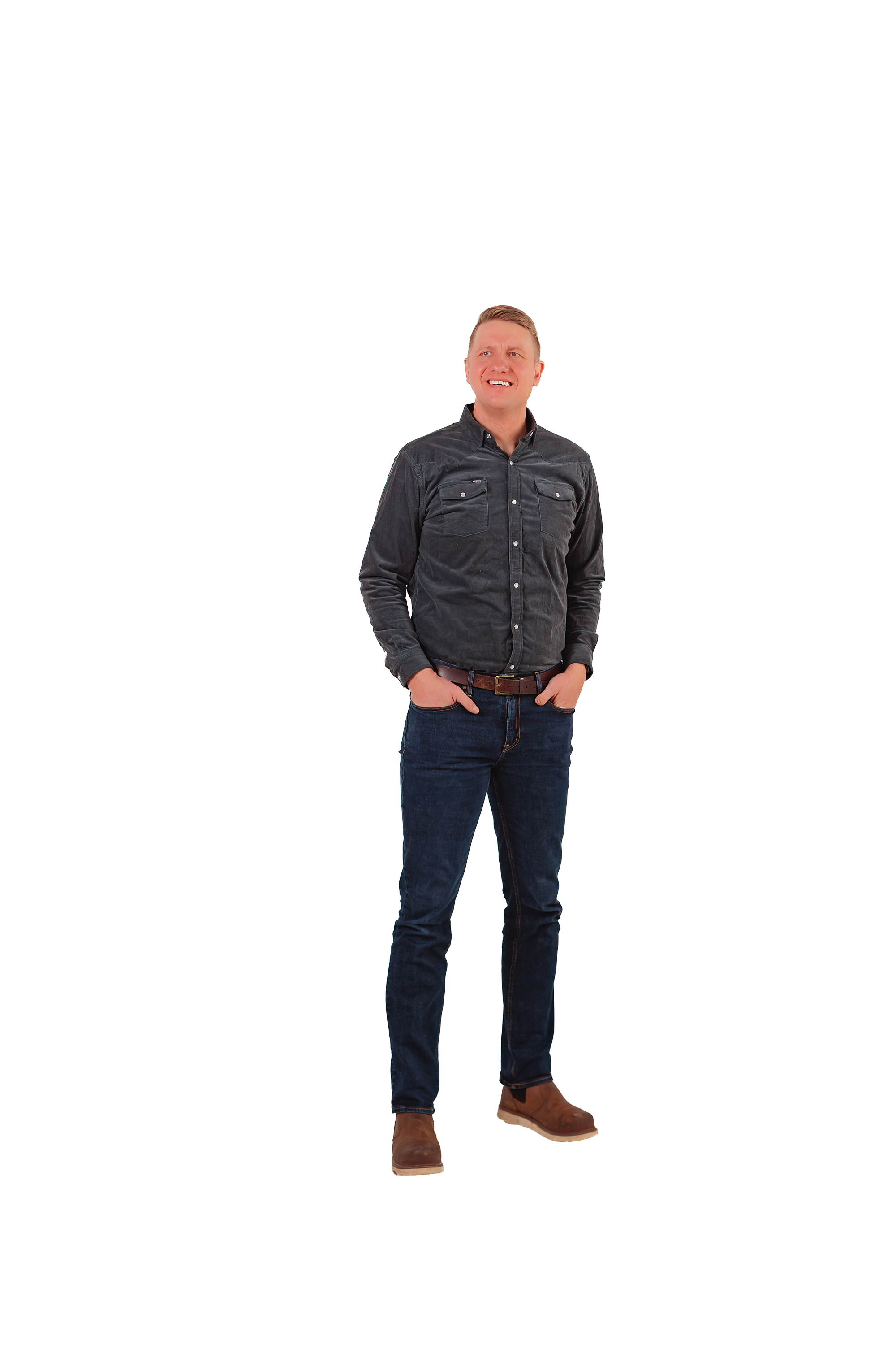 A man standing with his hands in his pockets, wearing a dark button-up shirt, jeans, and brown boots, smiling and looking slightly upwards.