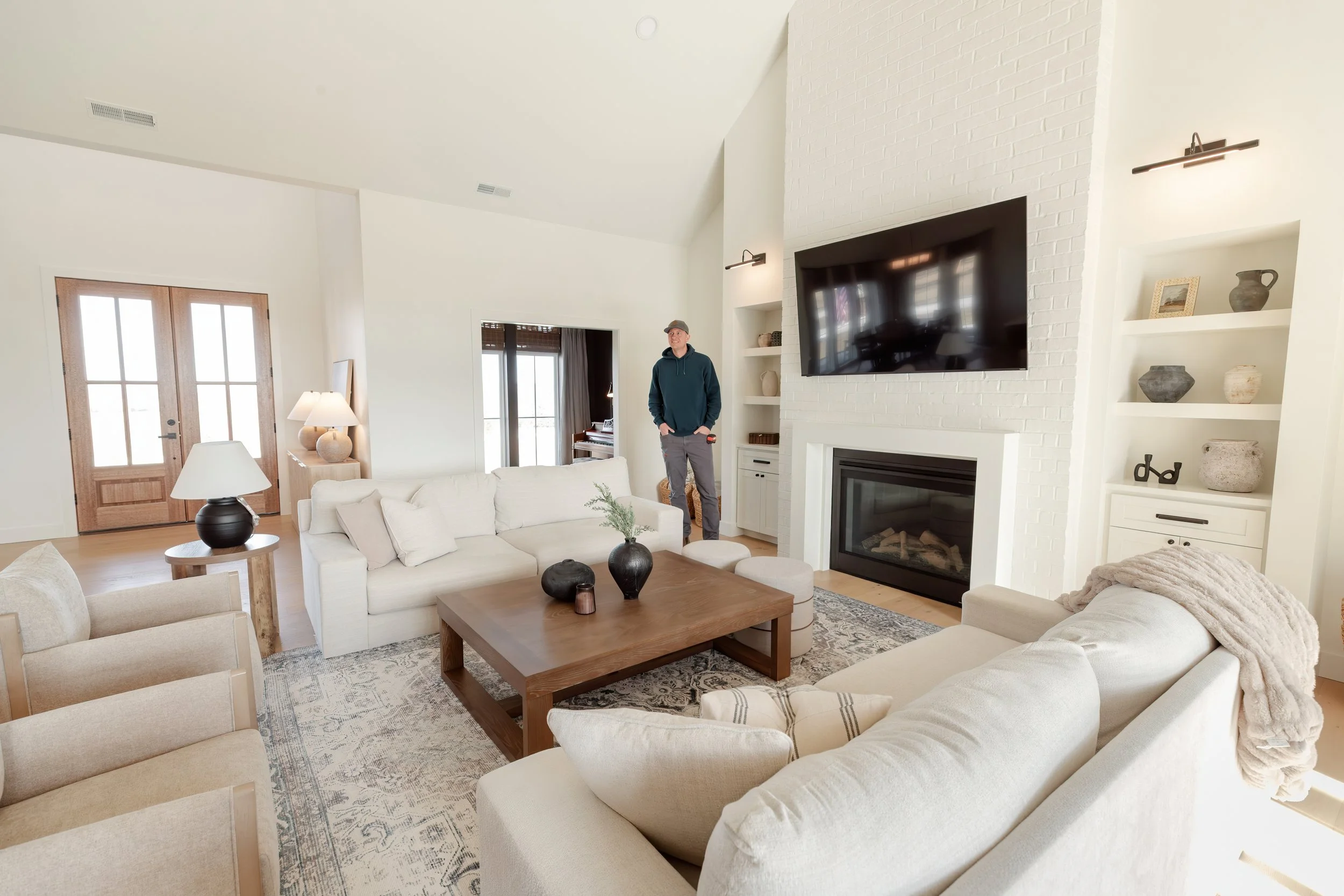 A spacious, modern living room with white sofas, a wooden coffee table, and a large flat-screen TV mounted on a white brick fireplace. There are built-in shelves with decorative vases, framed photos, and art. The room has wooden double doors and natural light coming through the windows, with a man in a blue hoodie and a gray cap standing near the fireplace.