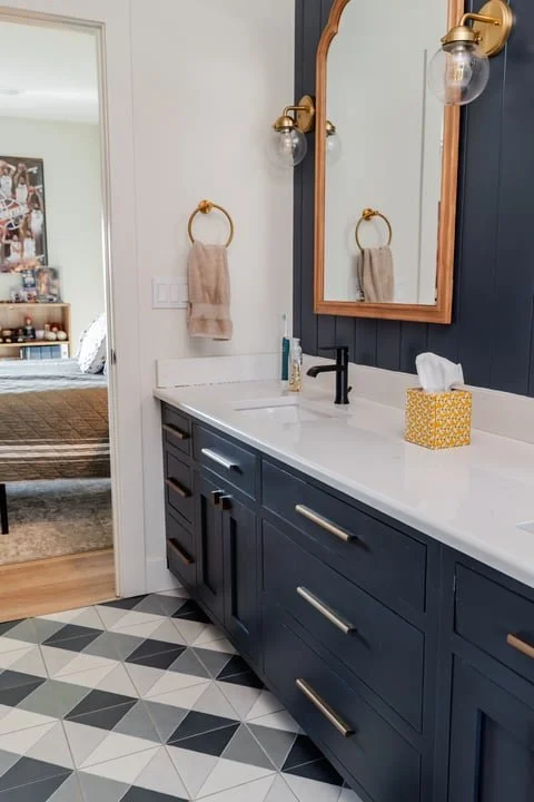 Modern bathroom with navy blue cabinets, white countertop, gold fixtures, and a mirror. A towel hangs on a gold ring, and a yellow tissue box is on the counter. The floor has black, white, and gray geometric tiles.