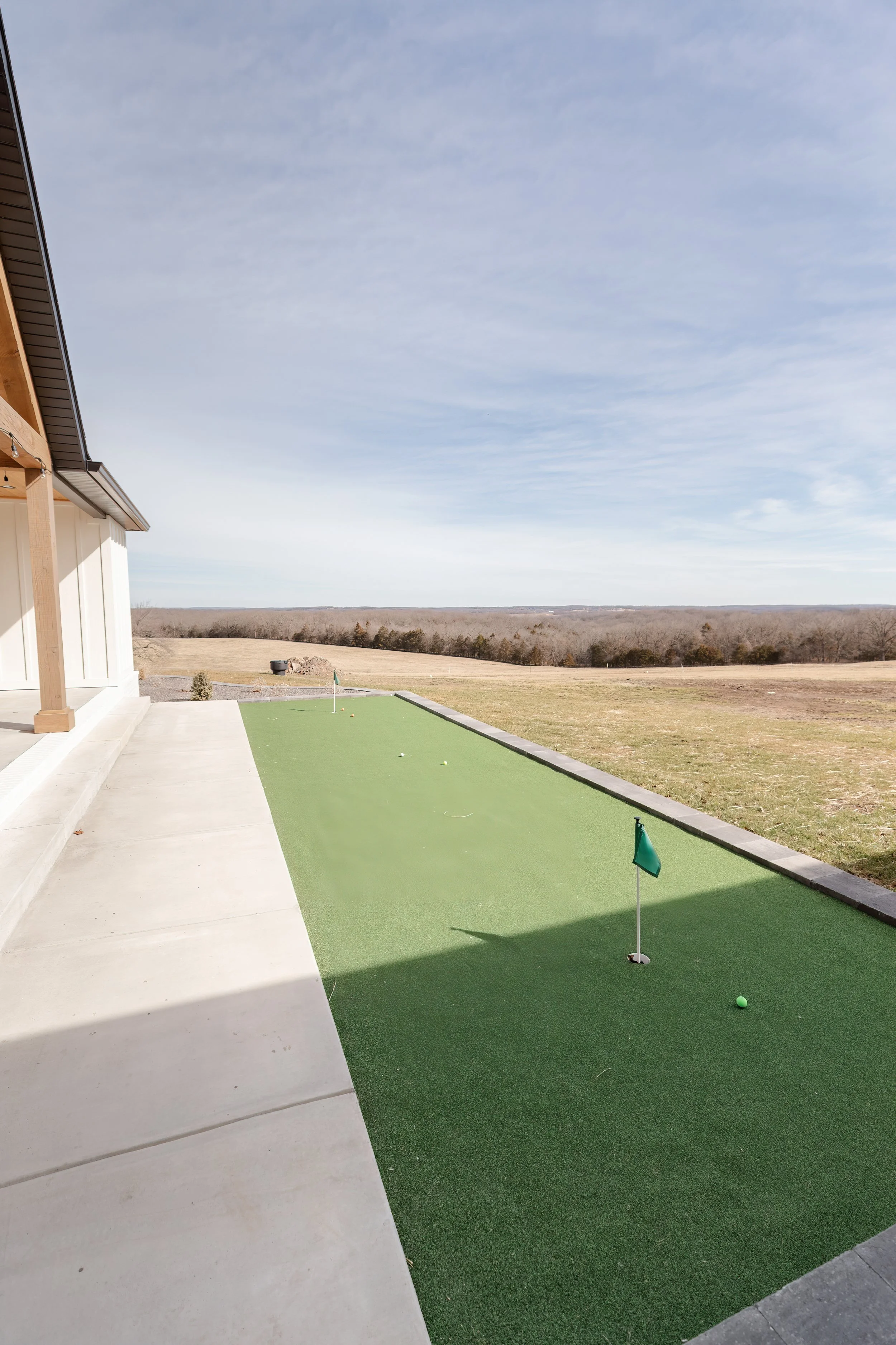 An outdoor mini golf putt-putt course with a green flag and ball on the artificial turf, next to a concrete pathway and a house with a wooden roof in a rural area with fields and trees in the distance.